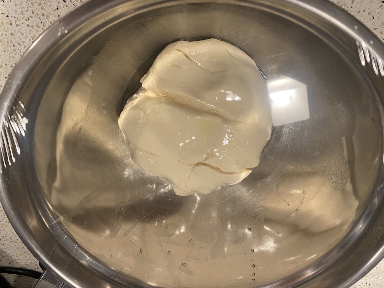 photo of a softball sized pizza dough ball in a steel bowl under plastic wrap, reflected off of the bottom and sides of the bowl to appear much larger