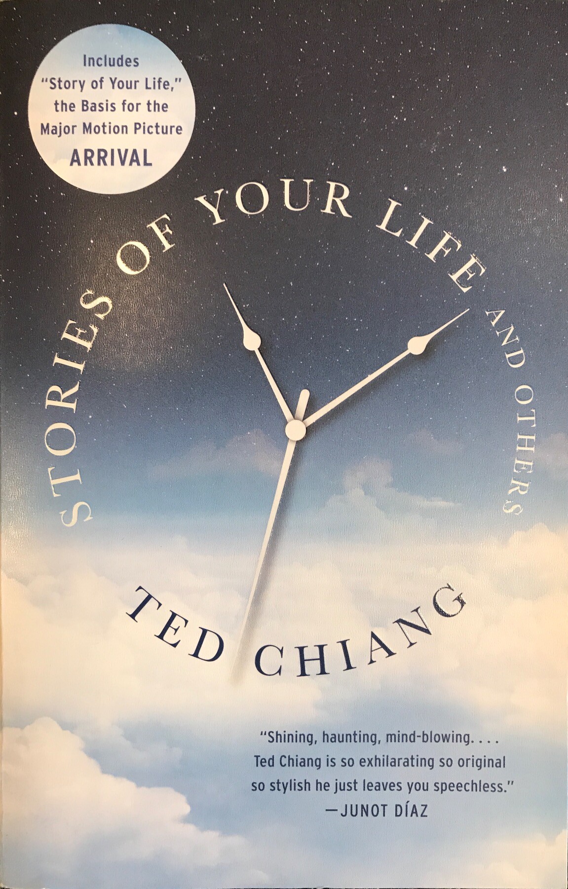 the current paperback cover of "Stories of your Life and others" by Ted Chiang. The title and author's name encircle the hands of a clock at about 11:10:30, over field of stars on the top half and cloudy sky on the bottom half. Inset in the top left is a cloud circle that proclaims that this book "Includes 'Story of Your Life," the Basis for the Major Motion Picture ARRIVAL.