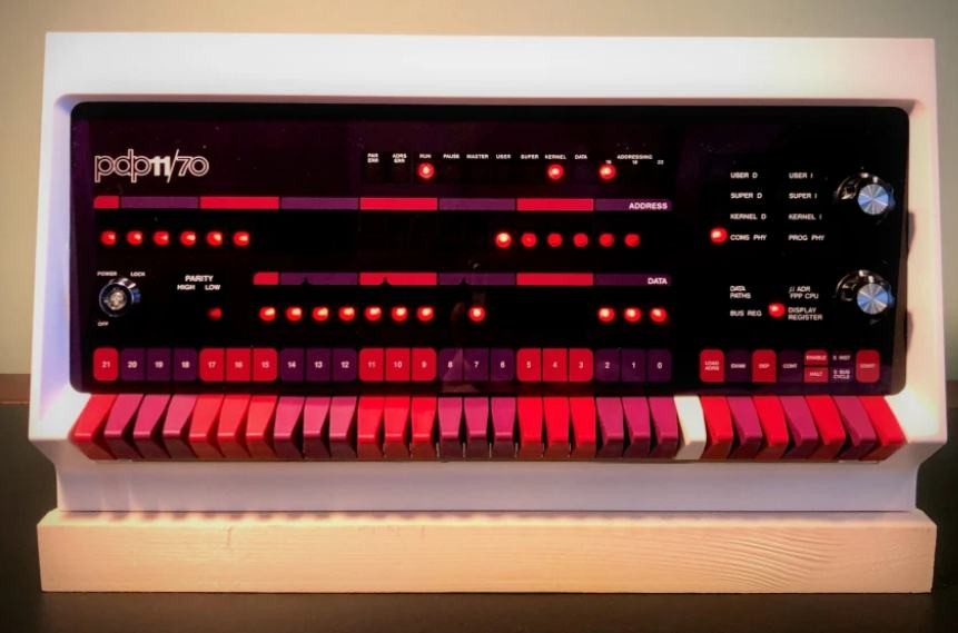 the front panel of a pdp-11/70 flush with rows of bright violet, fuchsia, and pink switches and buttons and red lights set on a black background over a wide row of violet, fuchsia, and pink keys in an off-white case