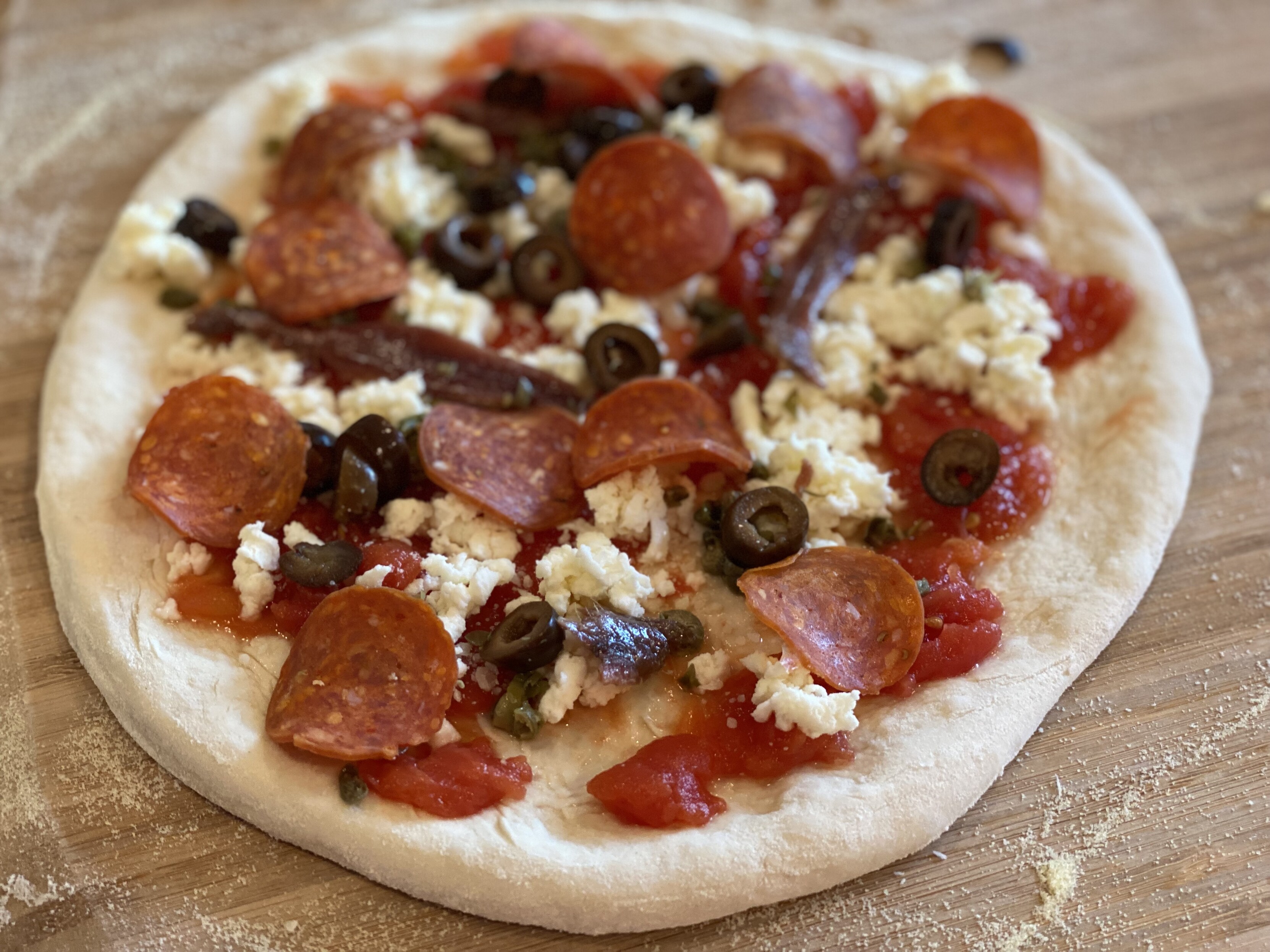 neapolitan pizza prior to baking, on the peel, topped with hand crushed san marzano, anchovies, capers, black olives, mozz, pecorino, pep, olive oil