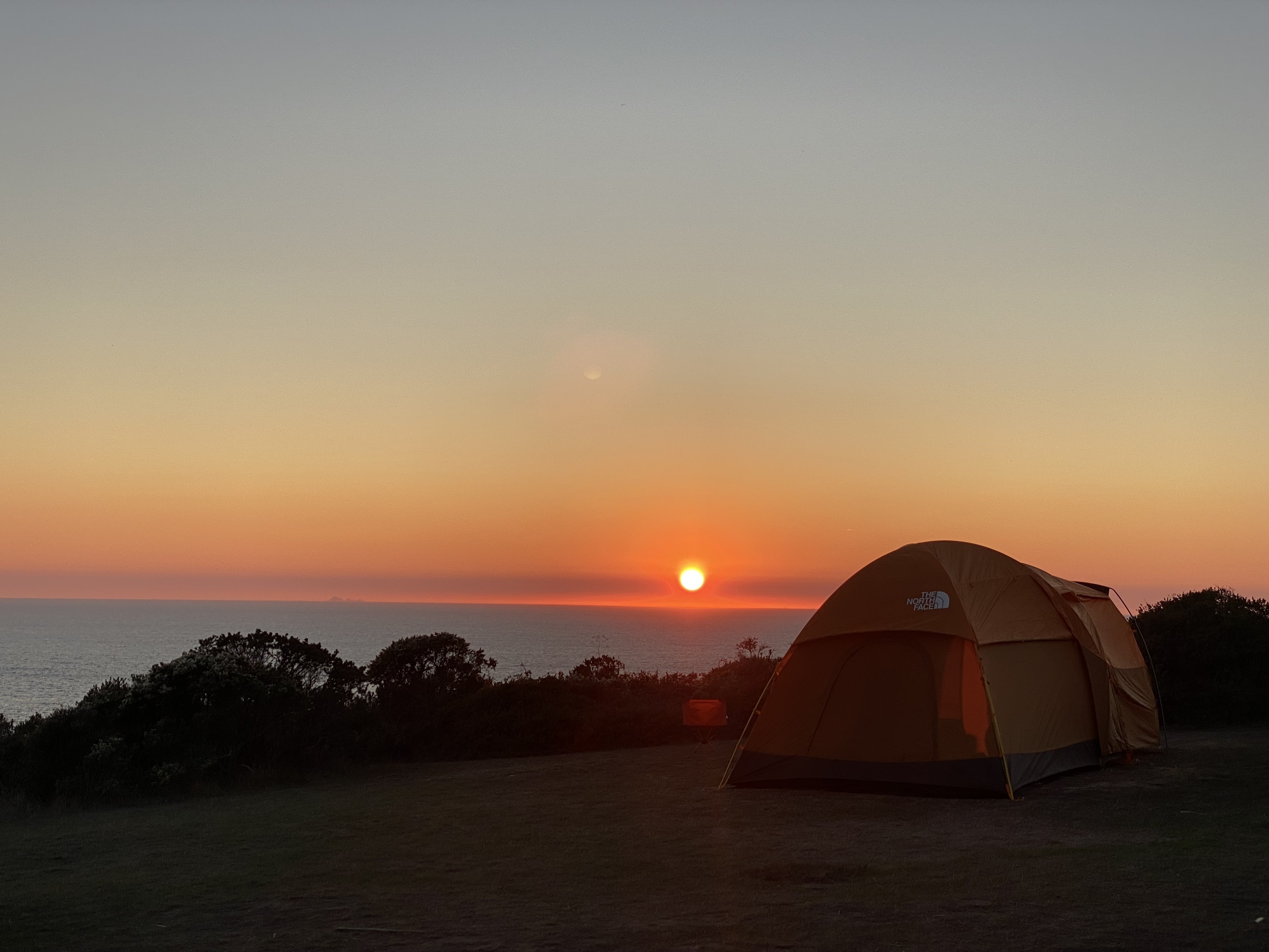 photograph of the sun setting over the pacific ocean, a line of bush, and a large orange tent in the foreground in a clearing