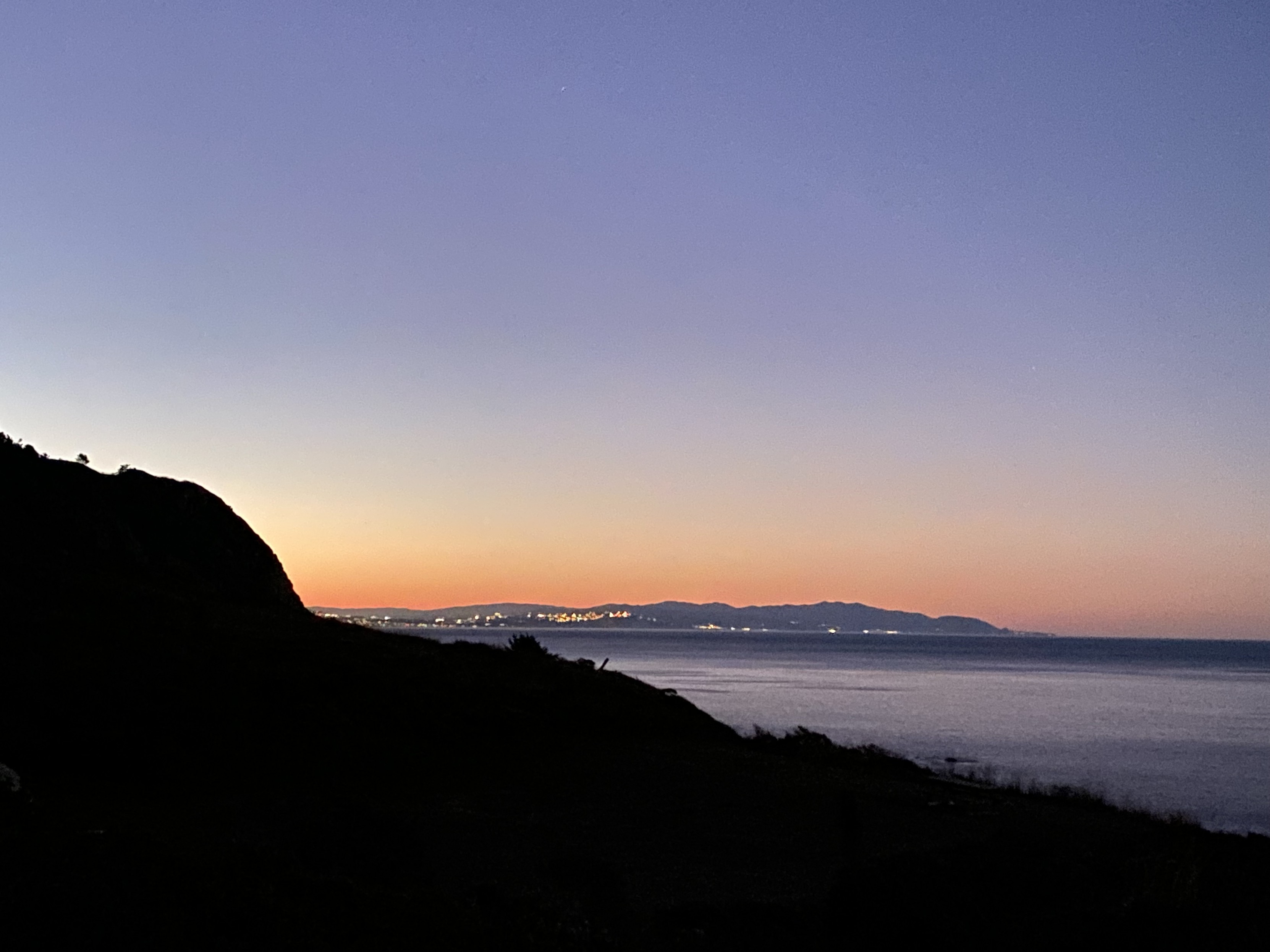 photo of the san francisco (or daly city or pacificia? I’m not sure) coastline from 20 miles north on the coast, looking south over a ridge as the sun set. urban lights are vibisle along the coast