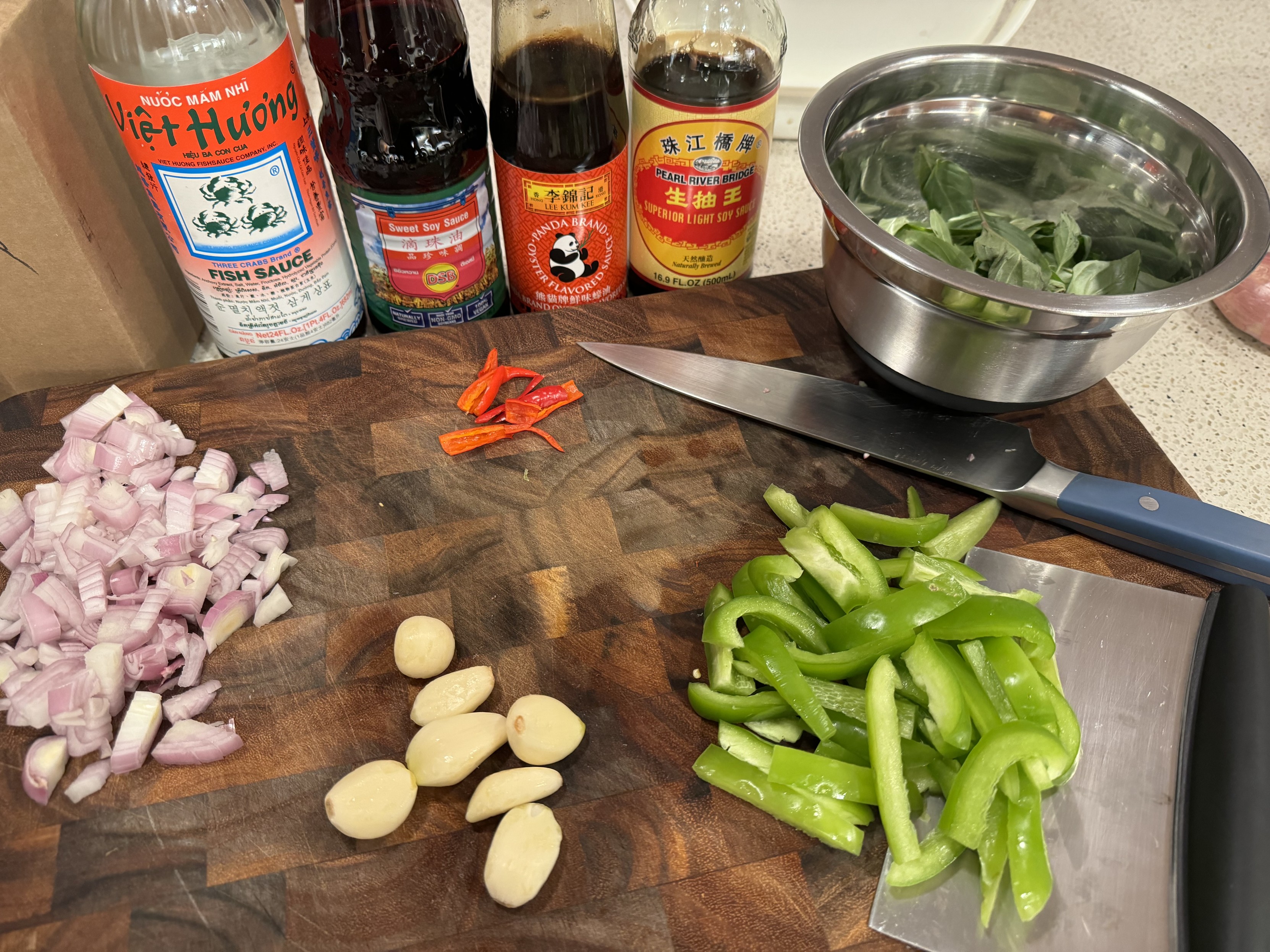 photo of cutting board with shallot, garlic, thai peppers, green pepper, and basil, bottles of sauce lined up to the rear