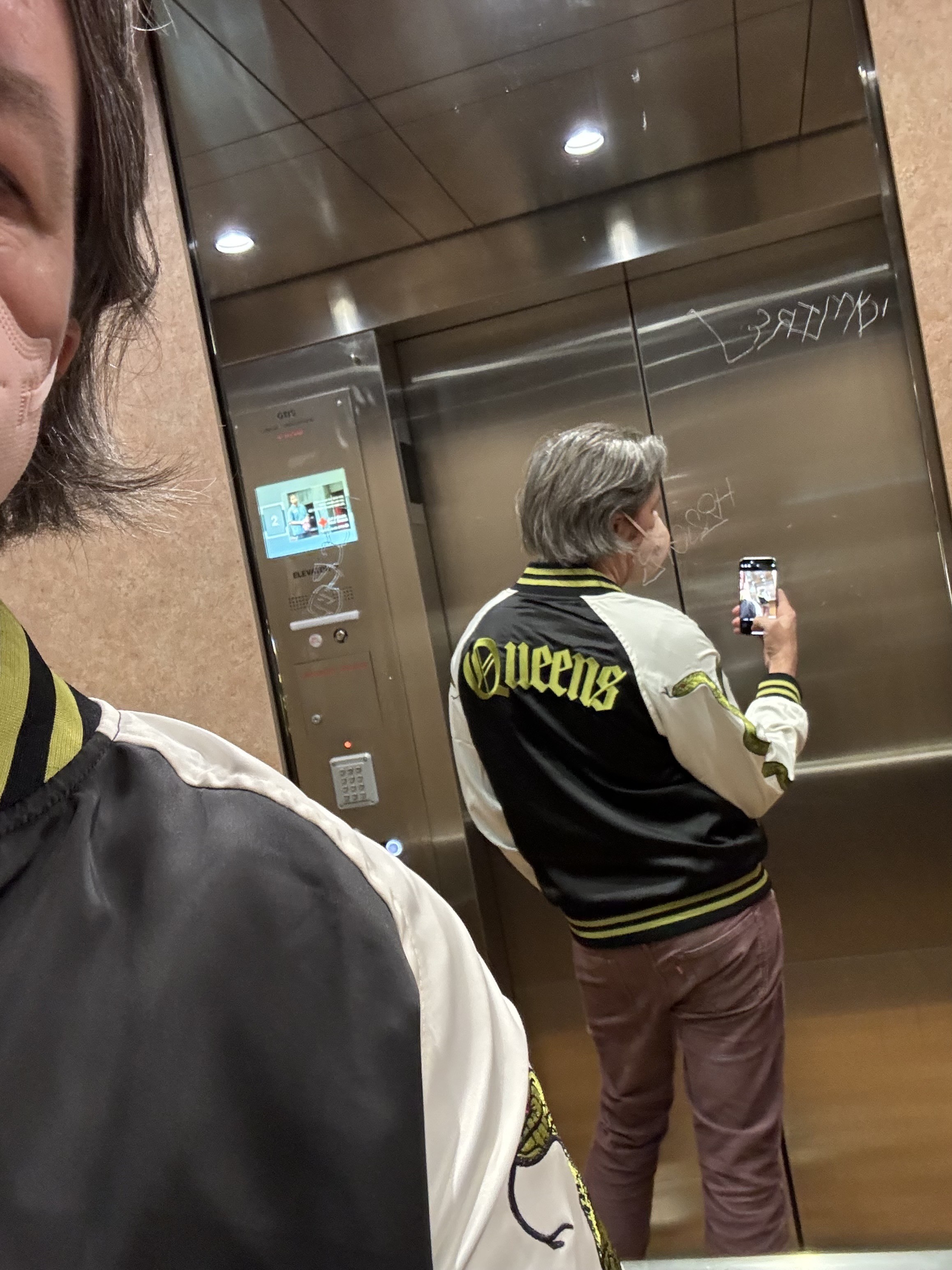 selfie in an elevator mirror wearing a new rock band jacket, black with white sleeves, green striped cuffs, collar, waist, and pockets, with a green two headed snake running up the sleeve, "Queens" stitched along the rear shoulder