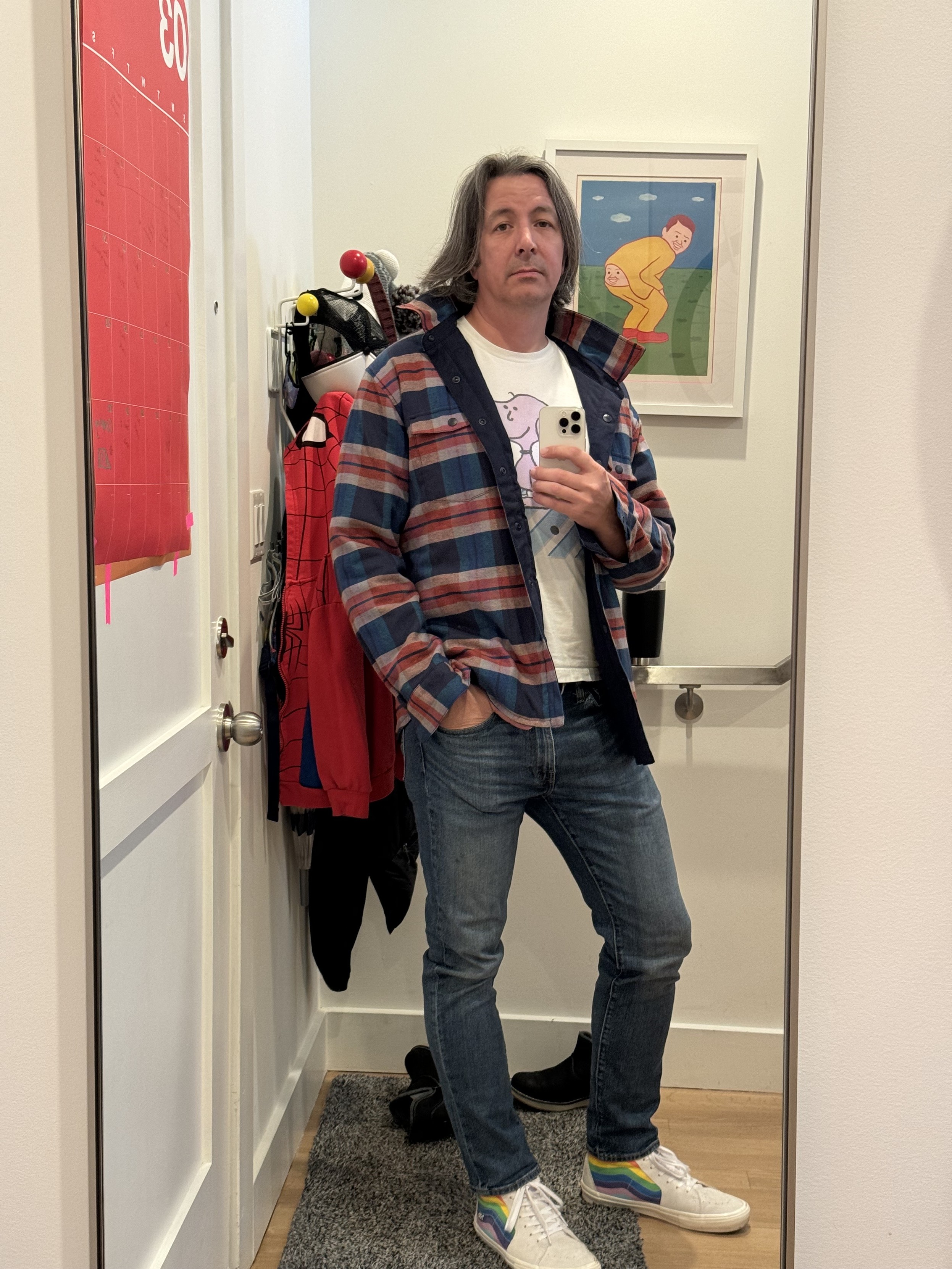 iphone selfie of me in our wall length mirror by the front door in a pink and blue flannel lined jacket over a sneppy t-shirt (fake snoopy), jeans, pride rainbow / white hi-top vans, in front of a joan cornella booty-poop print on the wall. there’s a huge red wall calendar for march taped to the door and an eames color-ball coat rack full of coats and masks next to me