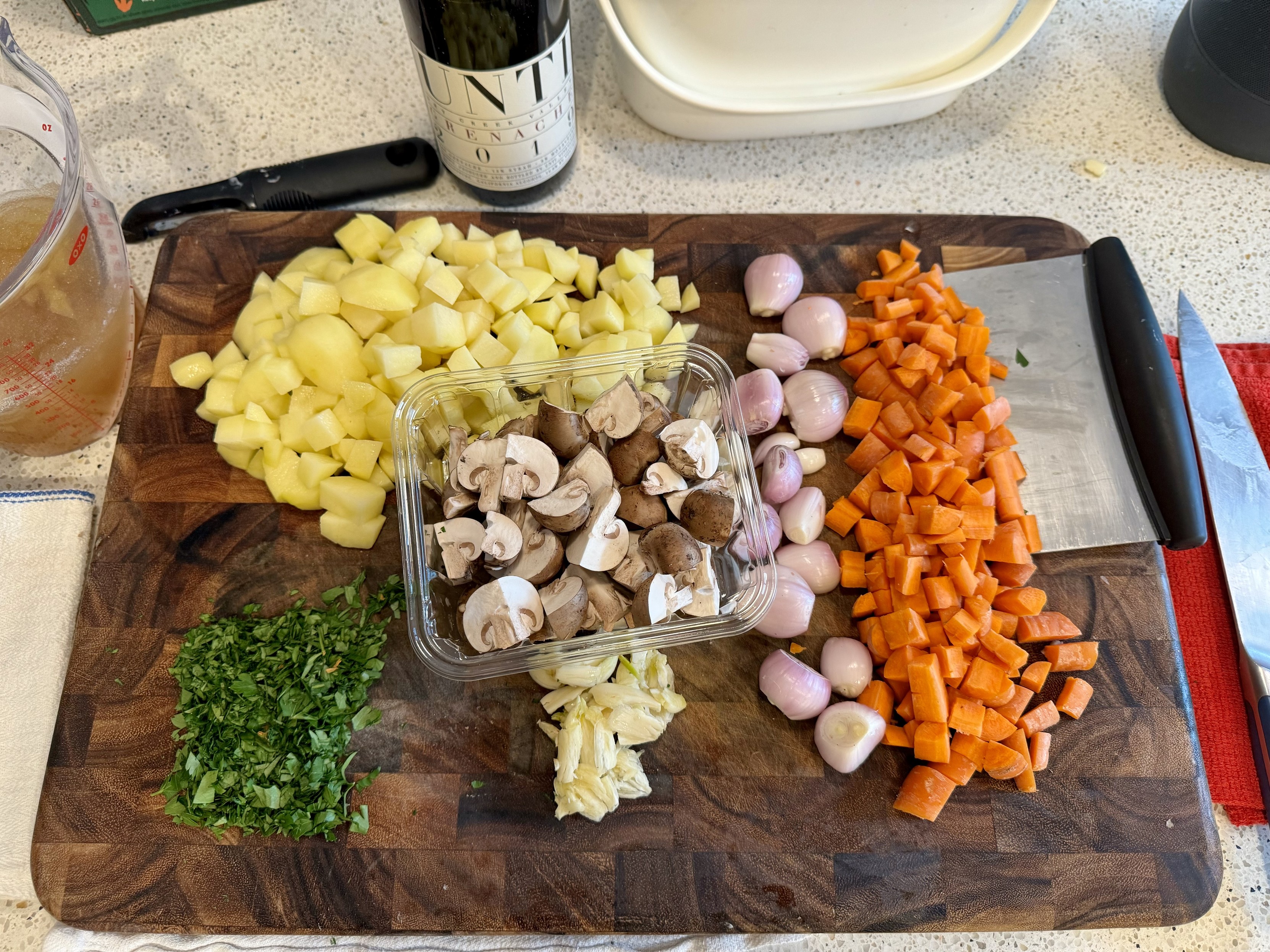 mise for coq au vin: a cutting board with diced carrots on top of a board scraper, quartered mushrooms, smashed garlic, peeled shallots, minced parsley, and diced potatoes (for a side). left to right behind the board: a few cups of broth with gelatin in a measuring cup, a peeler, a bottle of red wine, a compost container, ands a chef knife