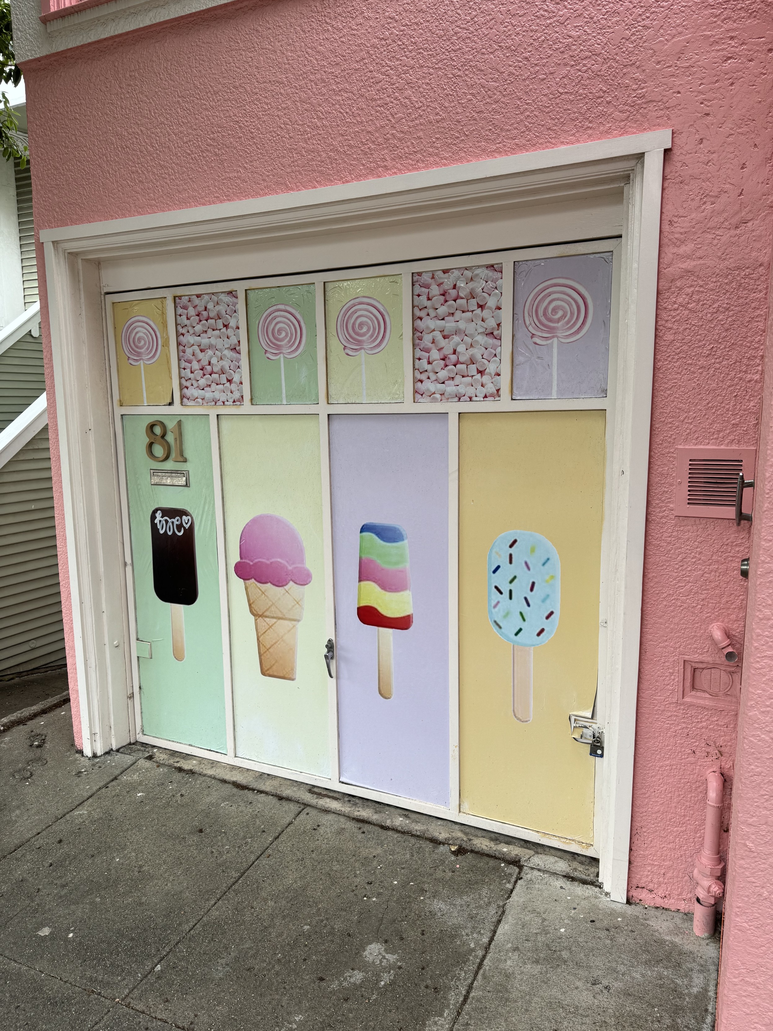 photograph of the garage door of a pink stucco house with white trimthe garage door panels are white with illustrations of an ice cream sugar cone, a fudgesicle, a rainbow sherbert pop on the main panels, and swirl lollys, and pastel mints on along the smaller top panelssomeone tagged the fudgesicle kinda clumsily in white spraypaint but at least used a heart in it so that’s nice I guess