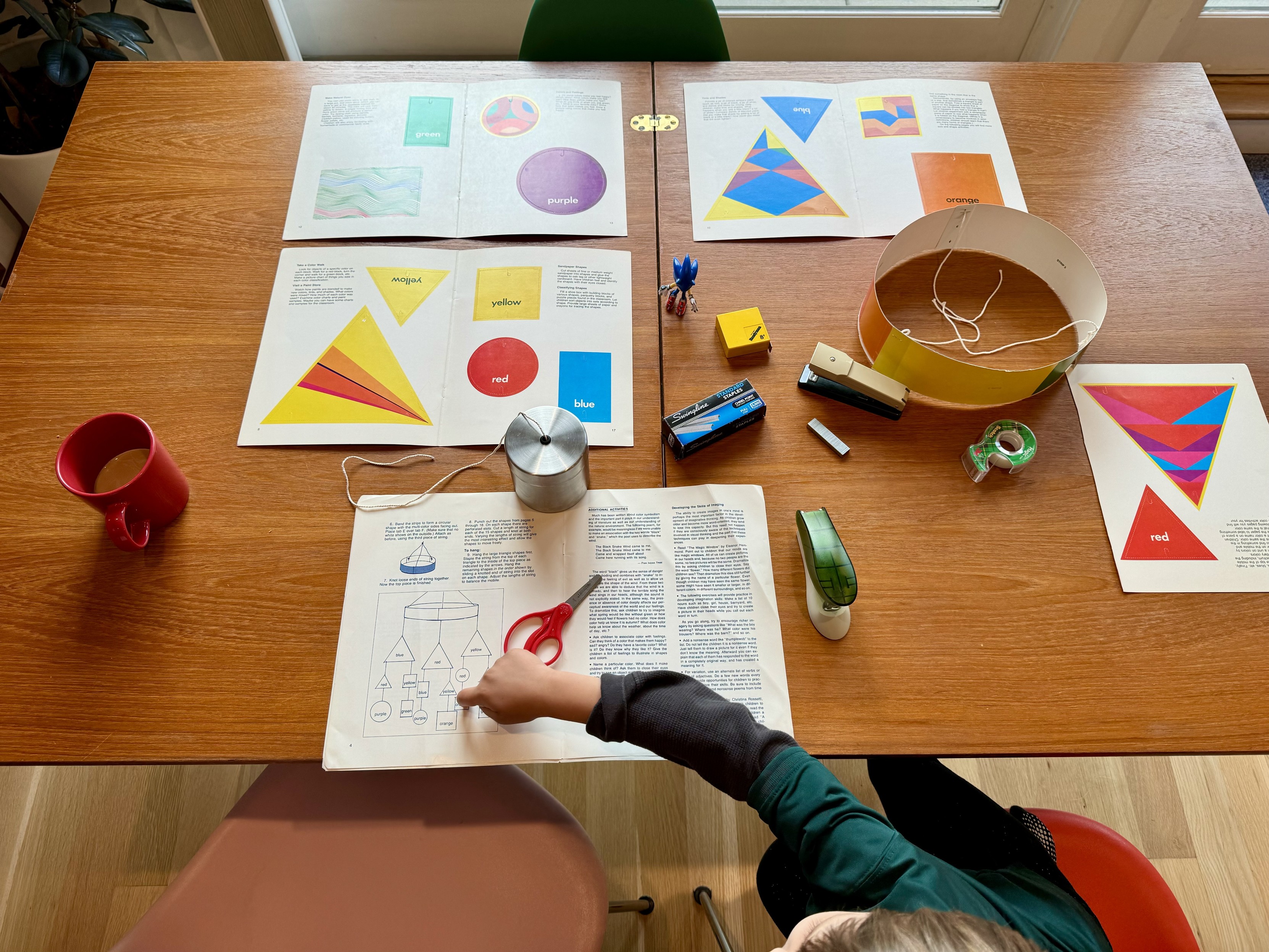 a photograph of the book’s staple binding removed and the eight color printed card stock pages spread as four landscapes two by two over the fifth containing instructions, set on our our dining table in rows of two, two and one, now jumbled with supplies including clear tape, two staplers, a box of staples, a measuring tape, and a spool of butcher’s twina sonic the hedgehog toy now supervises from the center of the tablethe color pages contain shapes in primary colors, perforated so as to be punched out, and a map of how to assemble them into a mobilethe hoop is assembled, and my kid is pointing at the map of next steps also visible are a few eames shell chairs in green, walnut, pink, and red,, a red cup of coffee, and a rubber plant in the corner