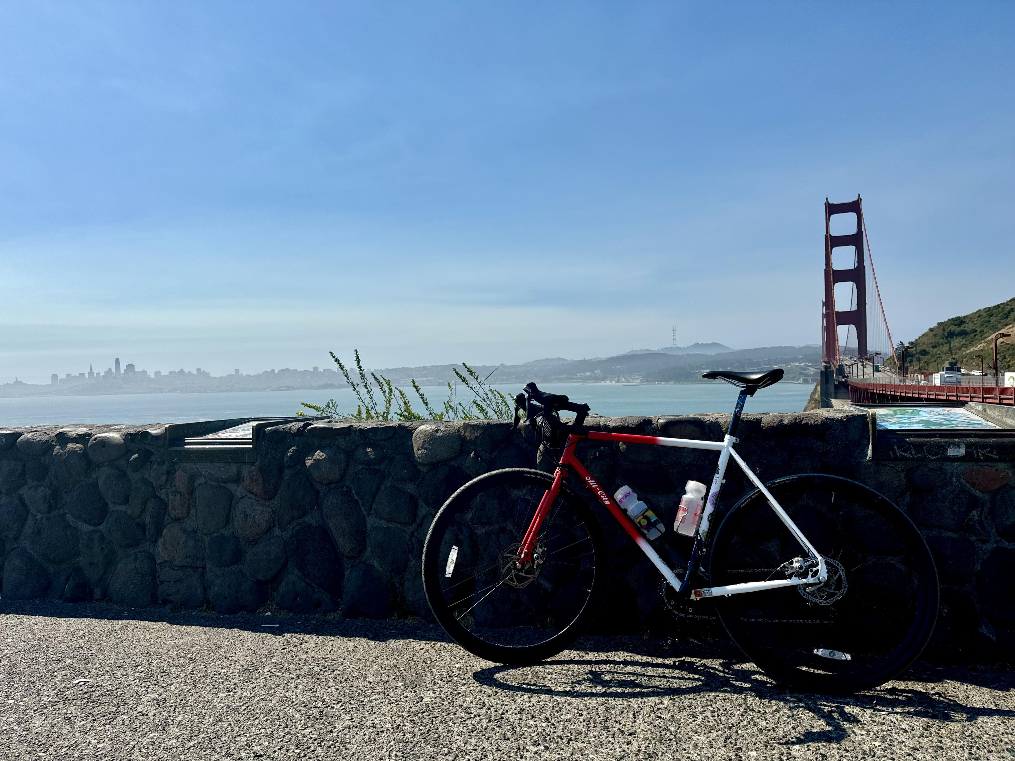 photo from the visitor’s lot on the north side of the golden gate bridge on a clear sunny day. the nearer, lower two-fifths of the photo contain a near stone wall, with my red and white steel frame gravel bike leaning against it on a gravel path. the distant, top three-fifths contain the san francisco bay, city skyline to the left, golden gate bridge to the right, and sutro tower in the distance