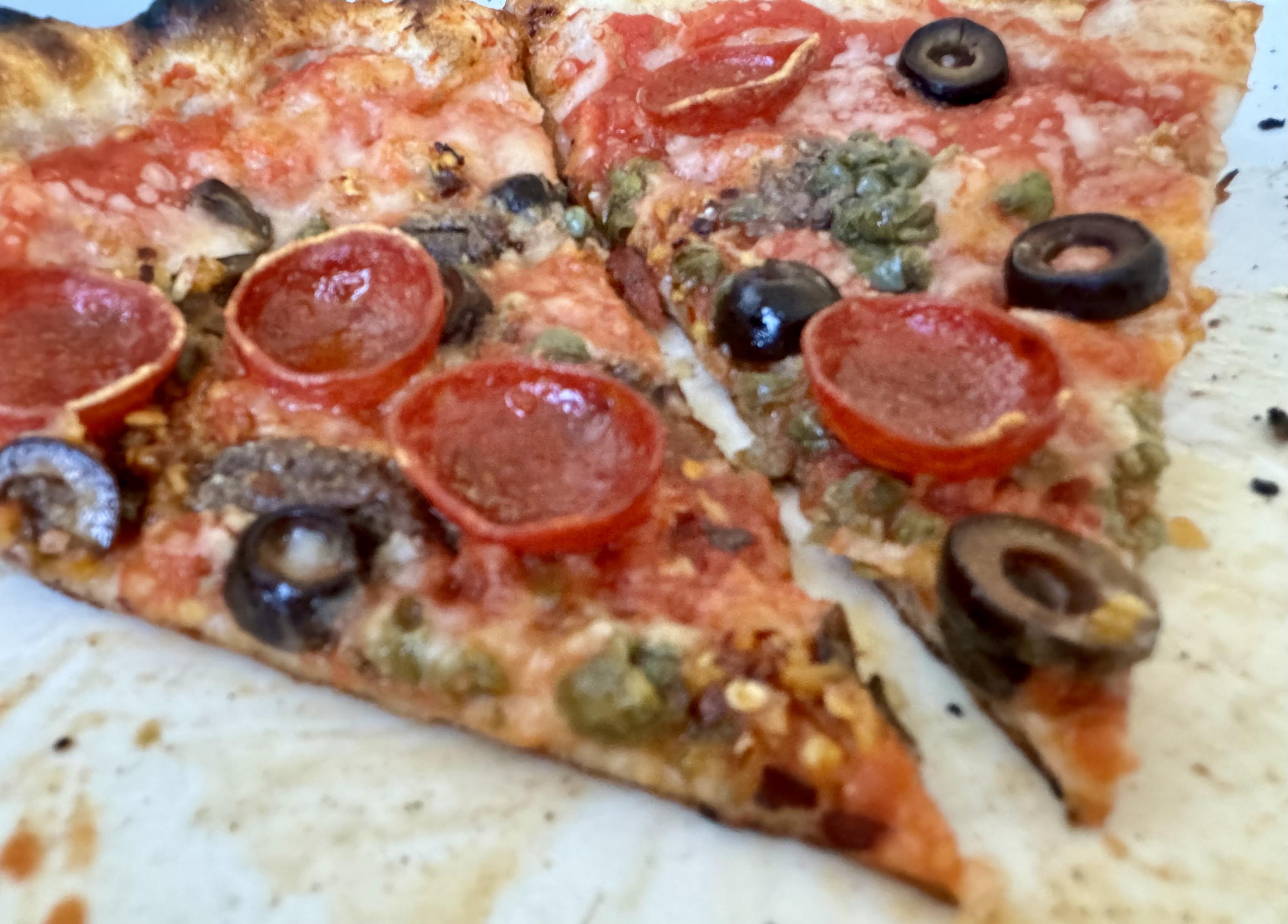 two slices of neapolitan pizza on a white plate topped with tomato sauce, pecorino romano, capers, black olives, anchovies, pepperoni, chili crisp, olive oil