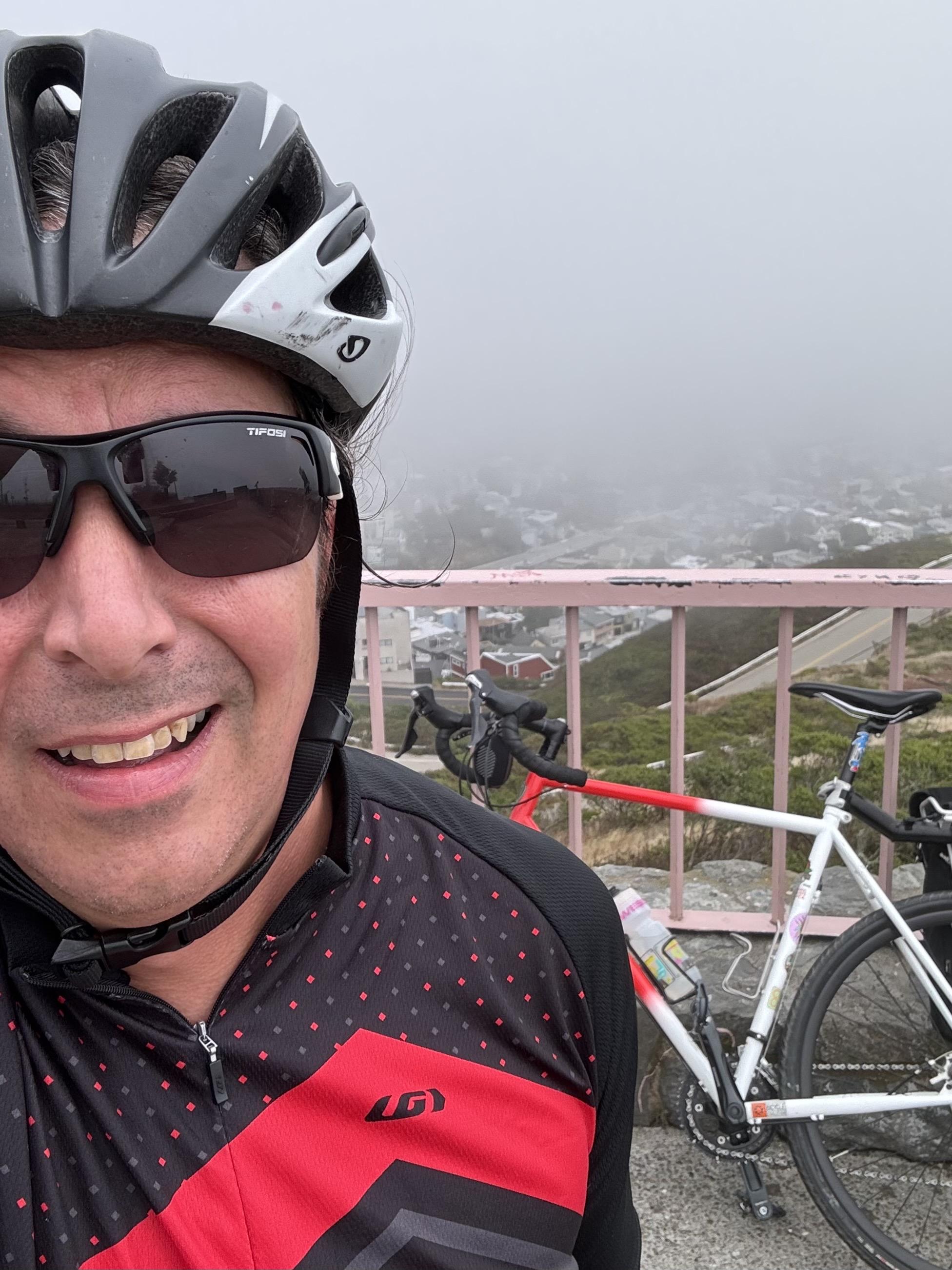 Selfie in a black and white giro helmet, transition glasses which are full tint black at this point, and a black cycling jesey with little red polka dots and a bright red zig zag across the front. My bike, a red and white gravel bike, is behind me, leaning up against the pink metal railing at Christmas Tree Point in San Francisco.