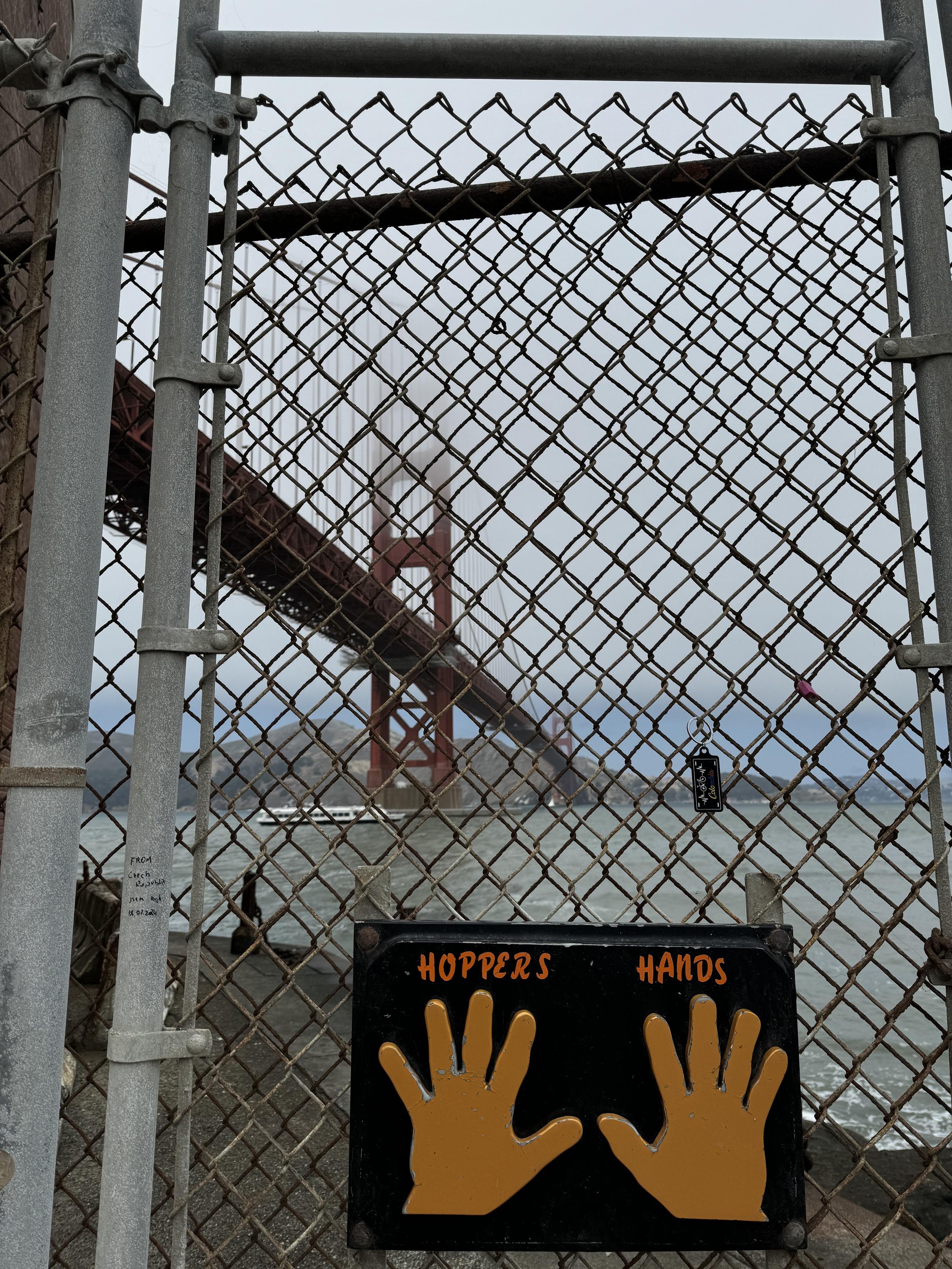 a photograph of the golden gate bridge, in san francisco, taken from below, in the parking lot of Fort Point, through a chain link fence. A black sign with yellow left and right hand prints at high-five height labeled HOPPERS HANDS is fixed to the fence as a touch point for people who walk, bike, hike, or whatever, to get here and then turn around as it is a dead end