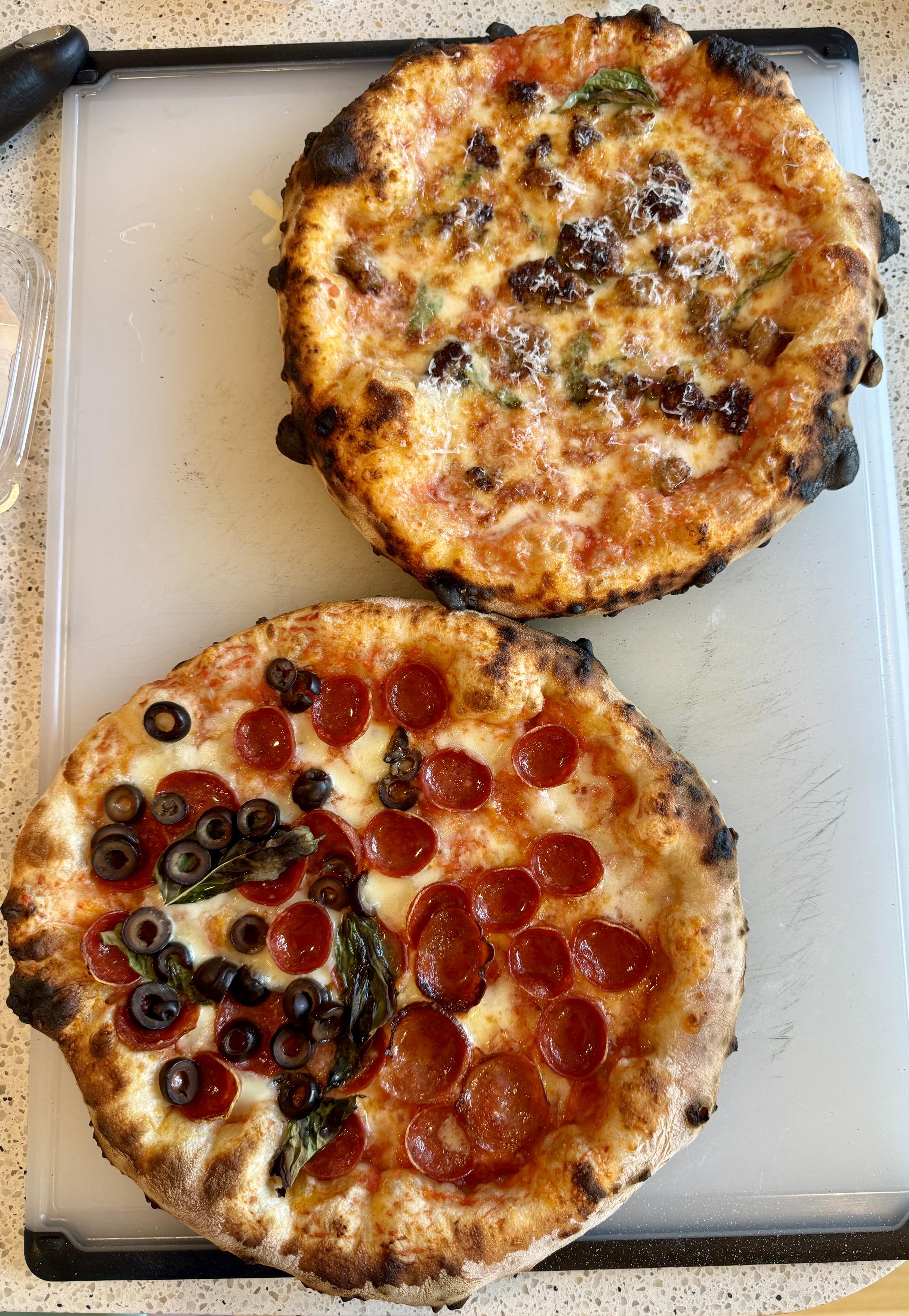 top down photo of the hugest oxo cutting board with two 12” neapolitan pizzas on it. top right is the sausage and cheese, bottom left is all pep, half olives