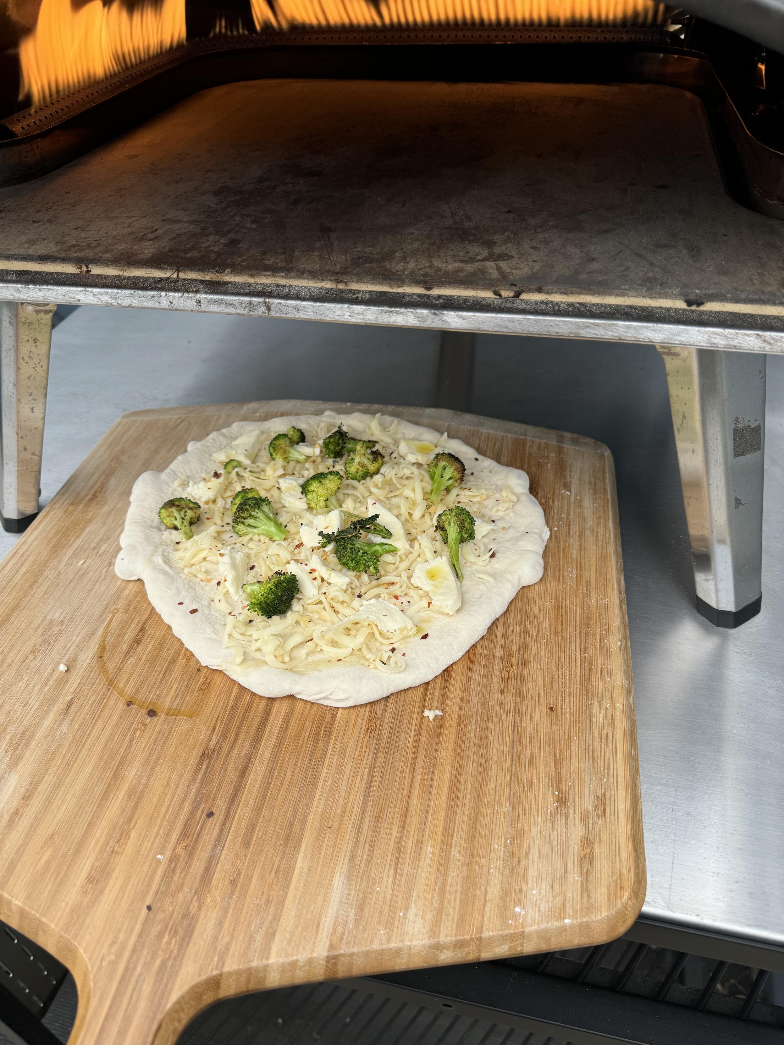 Photograph of the pizza before baking, topped with smoked provolone, mozz, red pepper flakes, grilled broccoli, and olive oil, on a bamboo peel resting on a stainless steel tabletop under an Ooni Koda 16 gas pizza oven. The flames of the oven are visible at the top of the photo as high as they get, and the oven is about 1000 degrees, the stone in the 800-900s, Fahrenheit. 