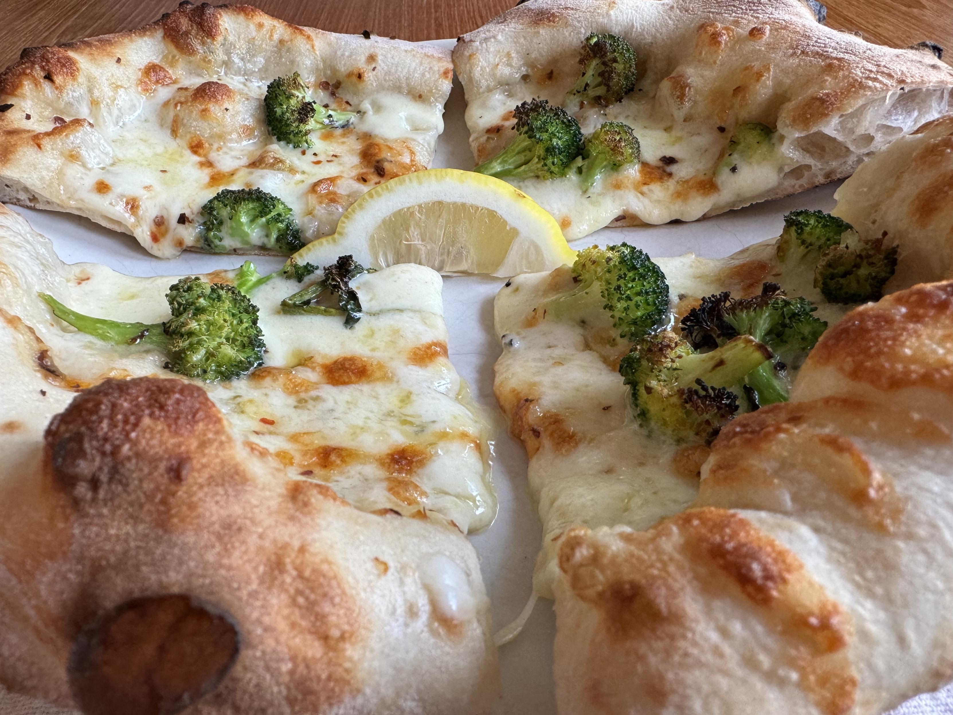 macro photograph of the pizza from inches above looking from one crust to the other, plated and sliced into quarters; the cheese has bubbled and browned in spots, the broccoli is charred a bit, and there’s a lemon wedge in the center, and the crust is browned with leopard spots