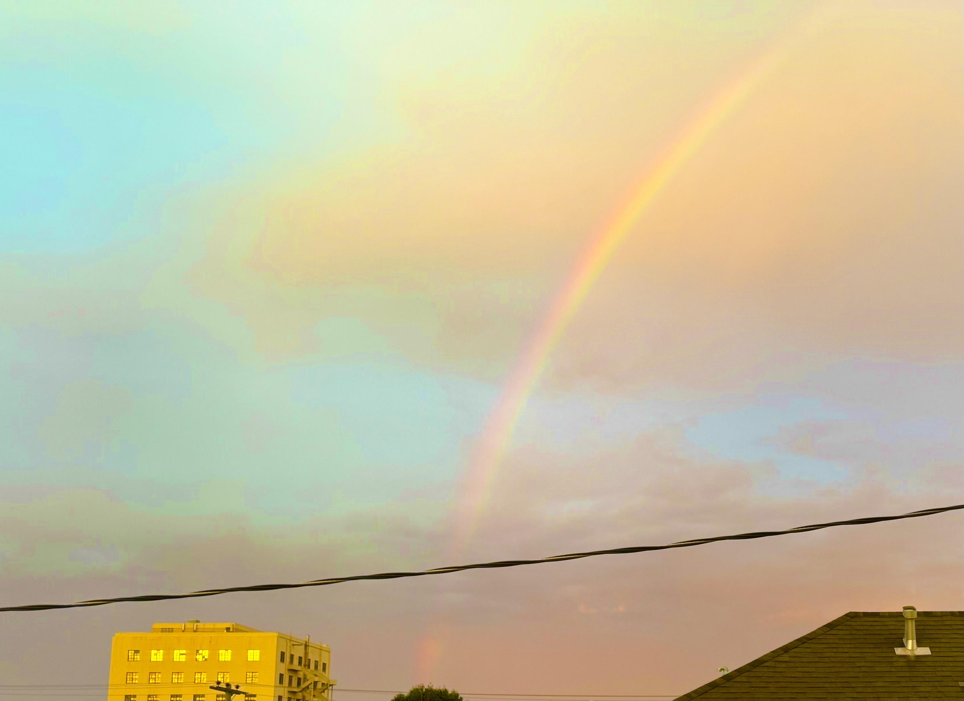 cropped photo of a rainbow over the mission in san francisco from the center bottom of the frame to the top 1/6th from the right-edge, a power line crossed 1/6th across the bottom edge and the top two stories of building in the bottom left face west and reflect the sunset in its windows. the sky is mostly cloudy with some blue