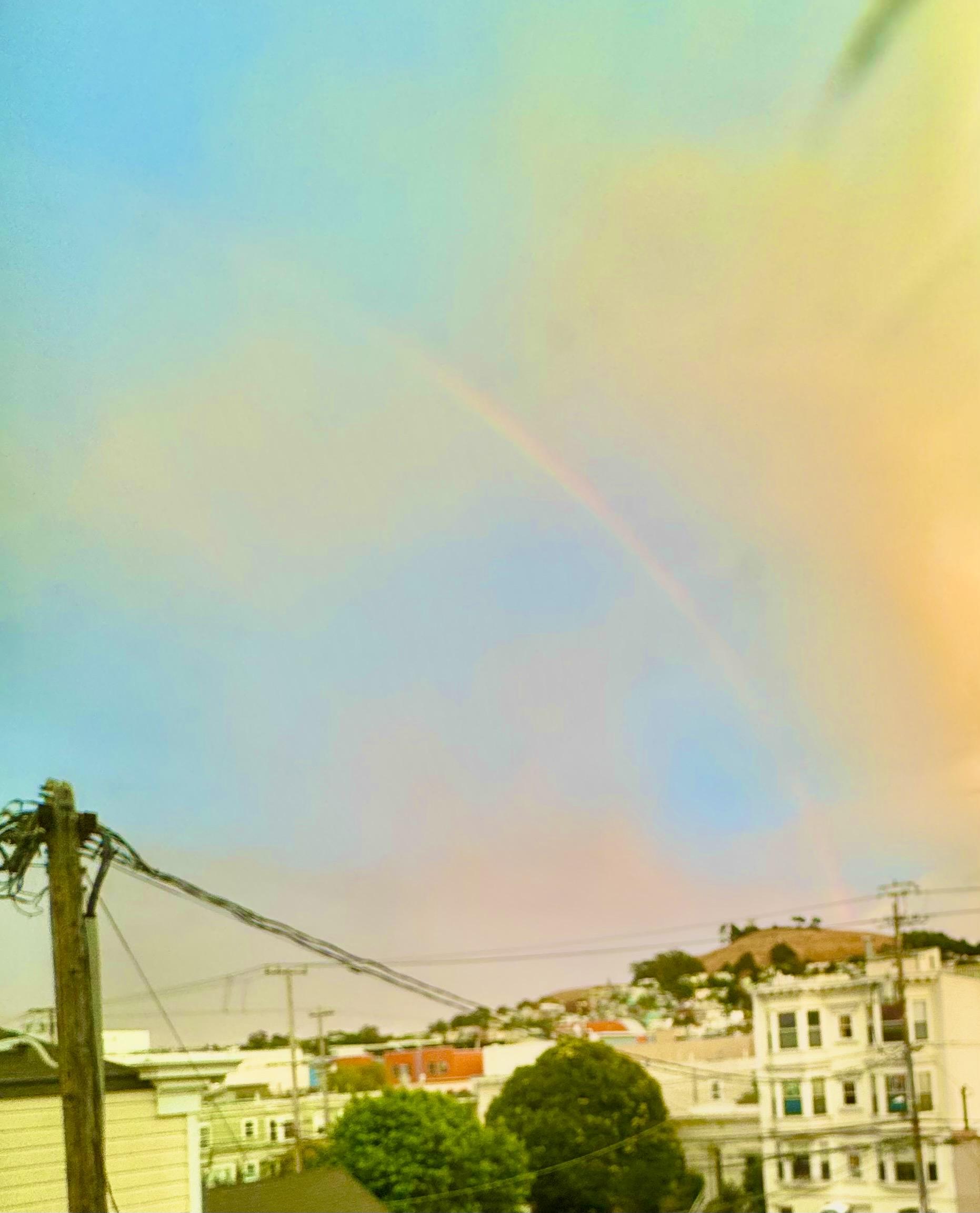 cropped photo of a rainbow touching down on the golden (dead) grass of bernal heights, the saturation is maxed out as is the luminance or something like that. A utility pole, trees, and buildings on 26th street fill the bottom 1/6th of the grame