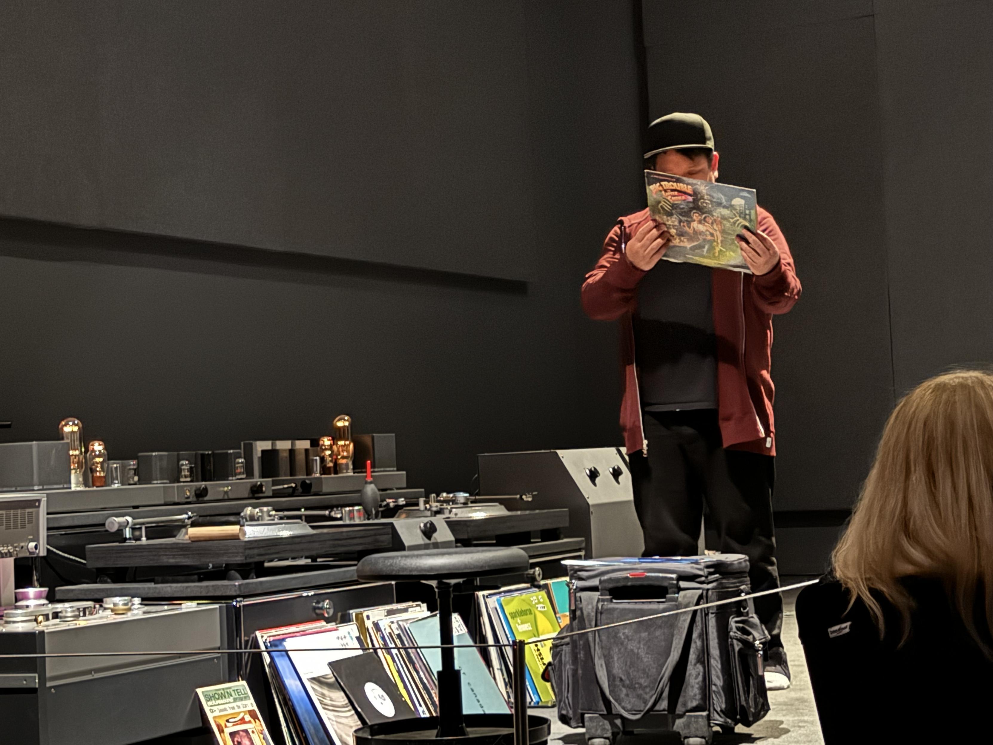 photograph of Kid Koala at SF MOMA's ‘Art of Noise’ exhibition in front of a knee-level table of fancy audio gear, with stacks of records, probably 60 or 80 or so, leaning against the table, and Kid Koala standing on front of it, looking at the back cover of John Carpenter’s soundtrack of Big Trouble Little China trying to decide what song to play for us