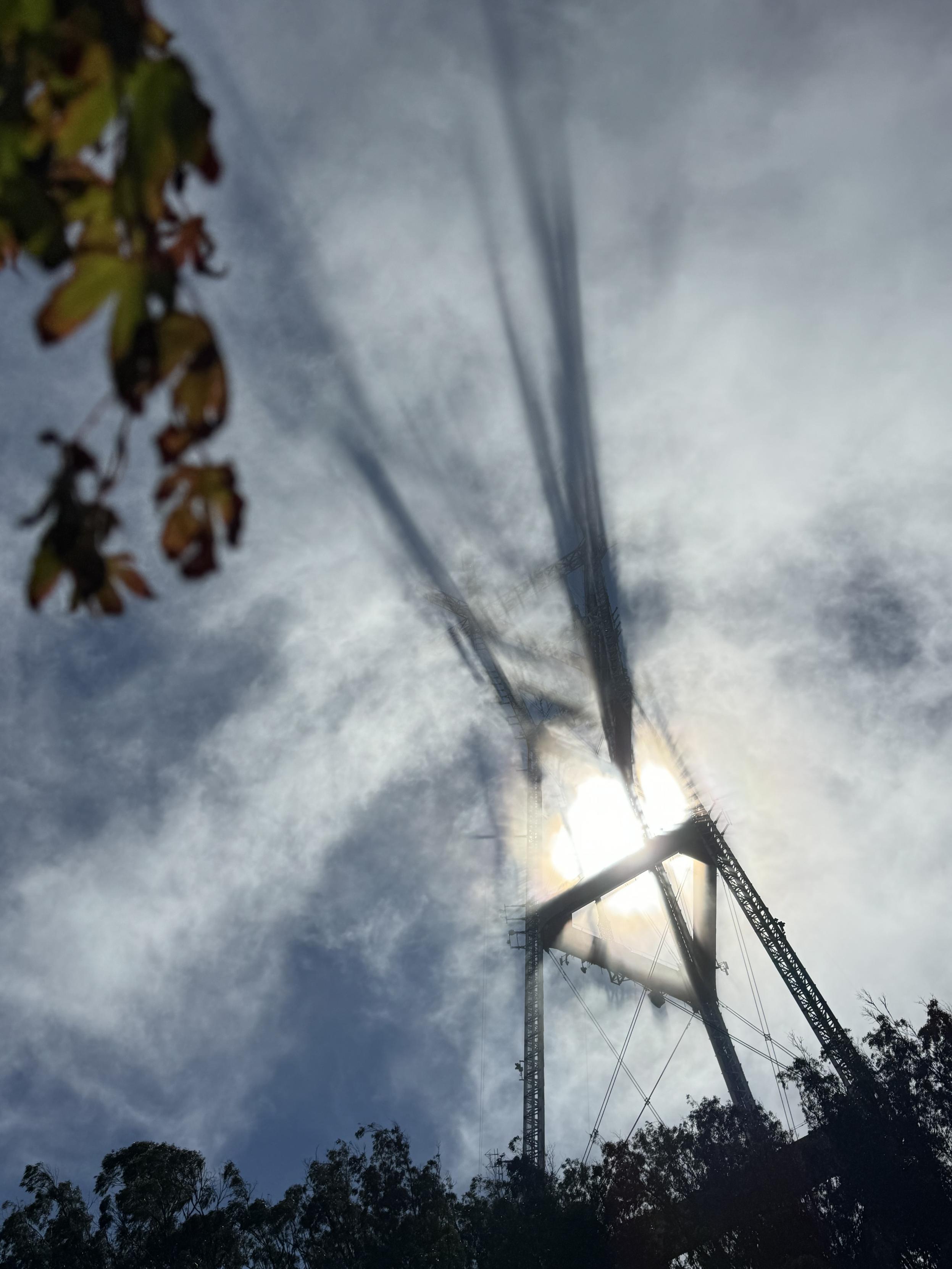 a photograph of a large radio tower, Sutro Tower, in san francisco at a dutch angle with the sun centered opposite, shining through the tower supports with fog throughout. the upper spires of the tower are casting long shadows in the fog