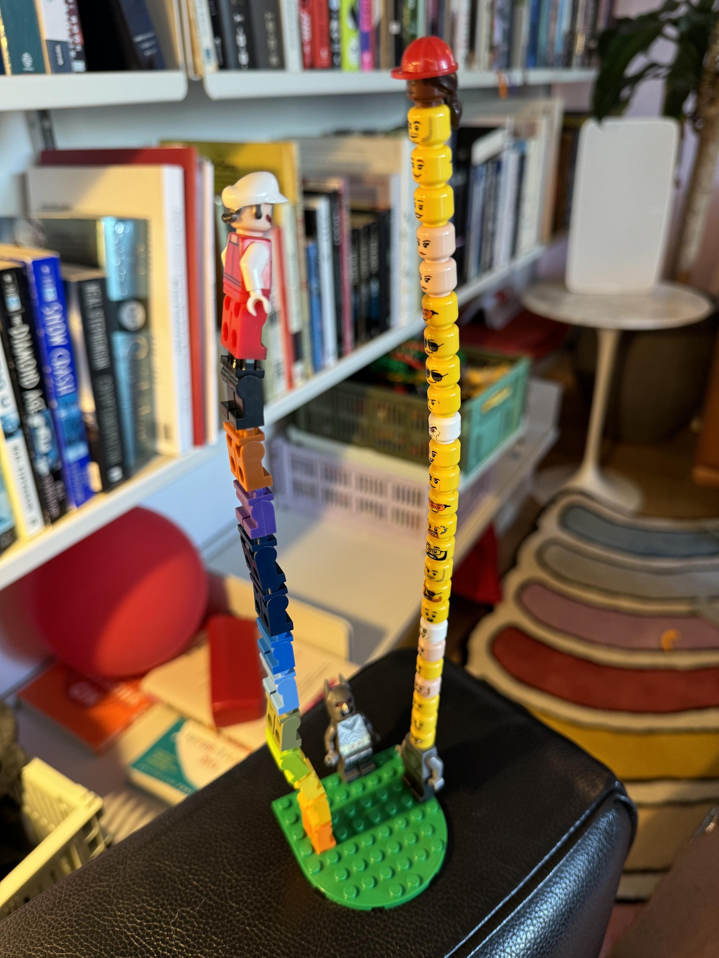 photograph of a stack of 10 lego legs or "pants" in a rainbow with fire mario on top standing on a small green platform facing a gray lego figure with like 20 heads stacked on top, the top one has long hair and is wearing a red hard hat. head-lord is taller than leg-lord by a few heads. a dumb old gray batman lego figure (normal height) stands between them looking up being mad about everything