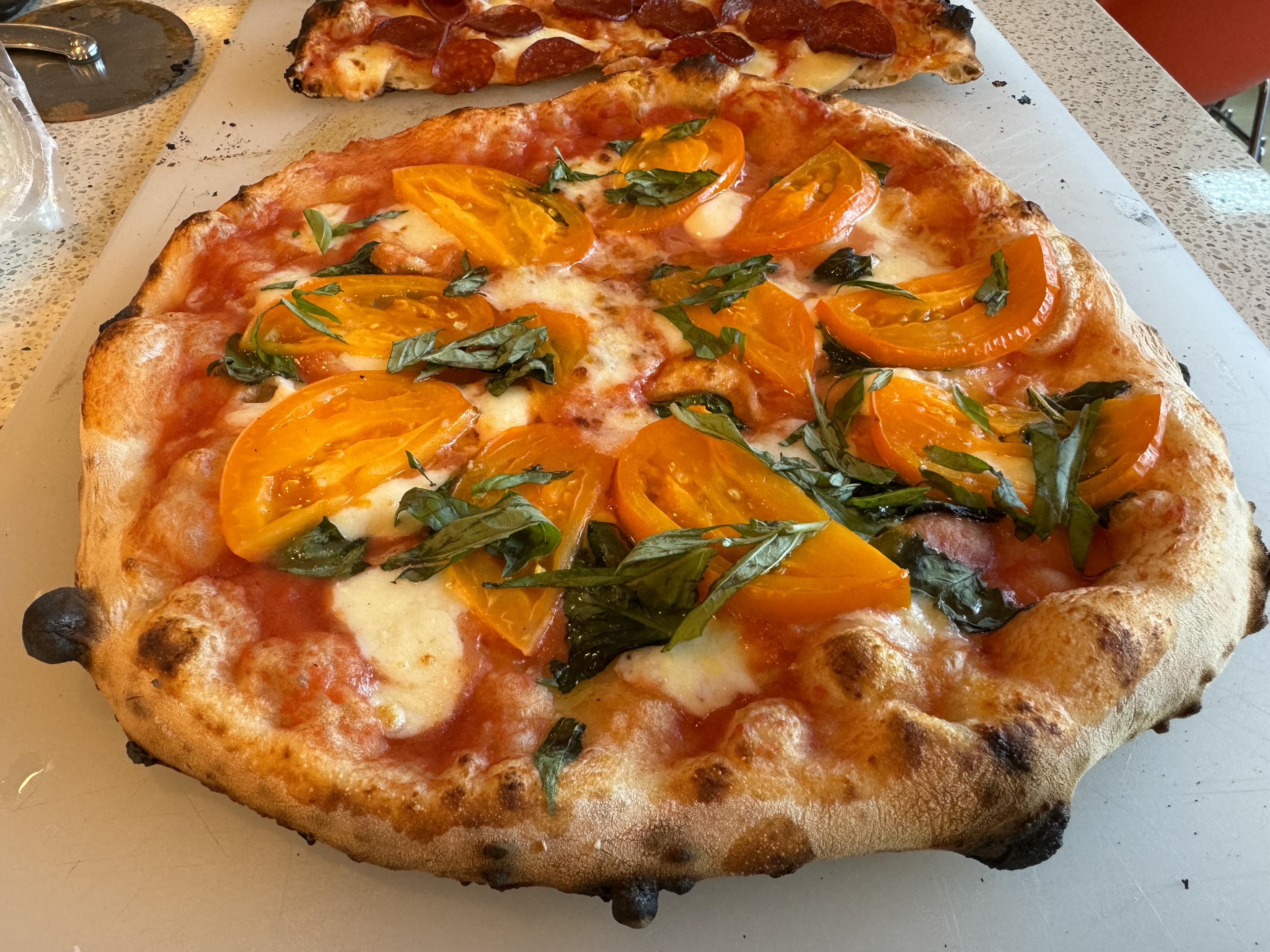 photograph of a margherita neapolitan pizza on a large cutting board, fresh out of the oventhe pizza has charred bubbles on the cornicione, and is topped with with tomato sauce, mozzarella, basil, and a fan-circle of thin, orange slices of a fresh garden tomato, topped with additional chiffonaded basil, and smoked sea saltin the background, also on the cutting board is half of a similar pepperoni pizza