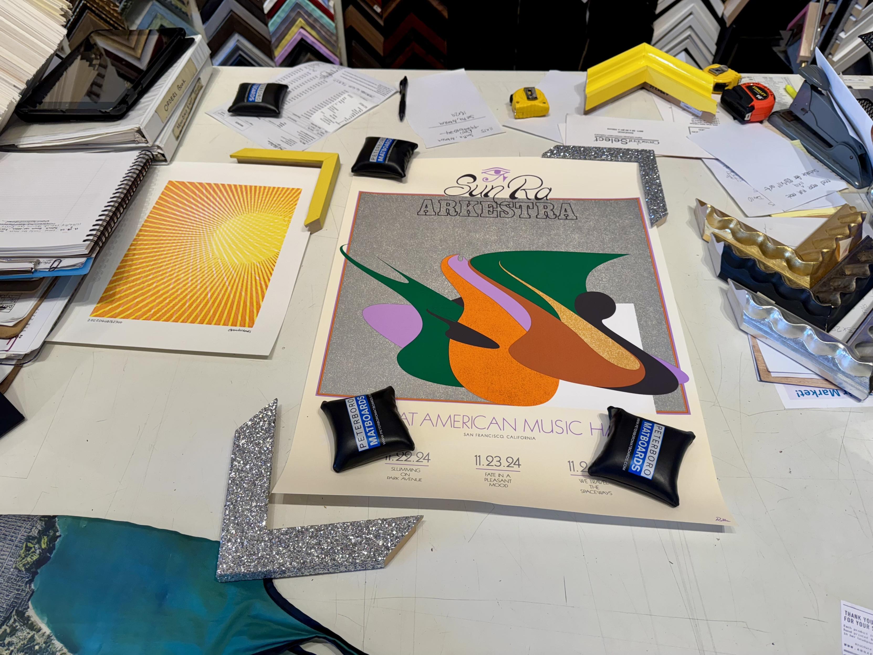 a pen plog on paper, and a larger poster, on a work table at the frame shop on the left is a medium sized yellow and orange pen line plot (done on a computer plotter) of a sun with rays casting outon the right is a Sun Ra Arkestra poster illustration of abstract green, orange, brown, and pink shapes over a gray square that takes up most of the poster, which has a creamy white background