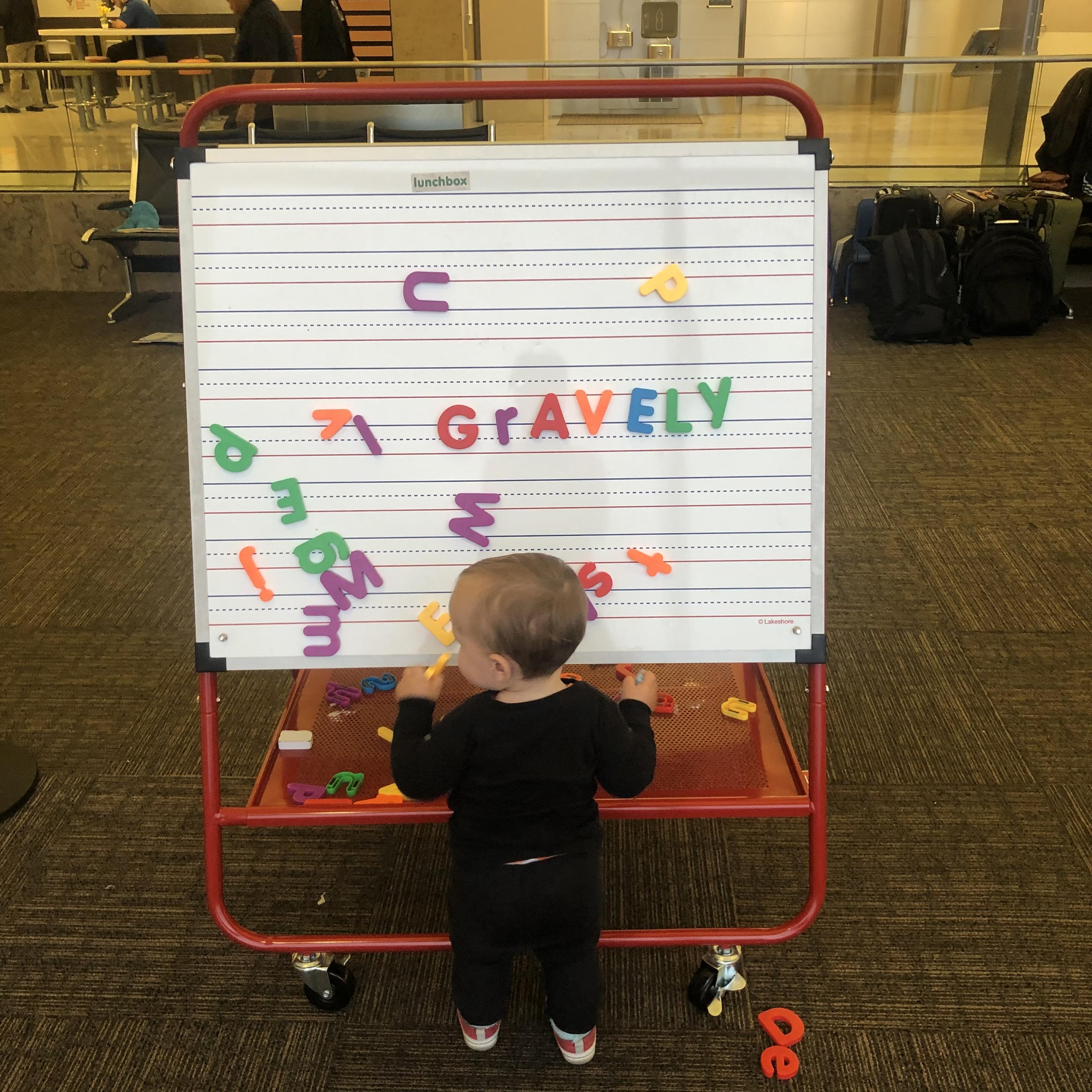 a toddler (my kid) under two years old wearing red shoes and black pants and a black shirt stands with their back to us in front of a giant magnetic easel with rainbow colored plastic magnetic letters scattered over it, including: G r A V E L Y across the middle in red, purple, red, orange, blue, and green