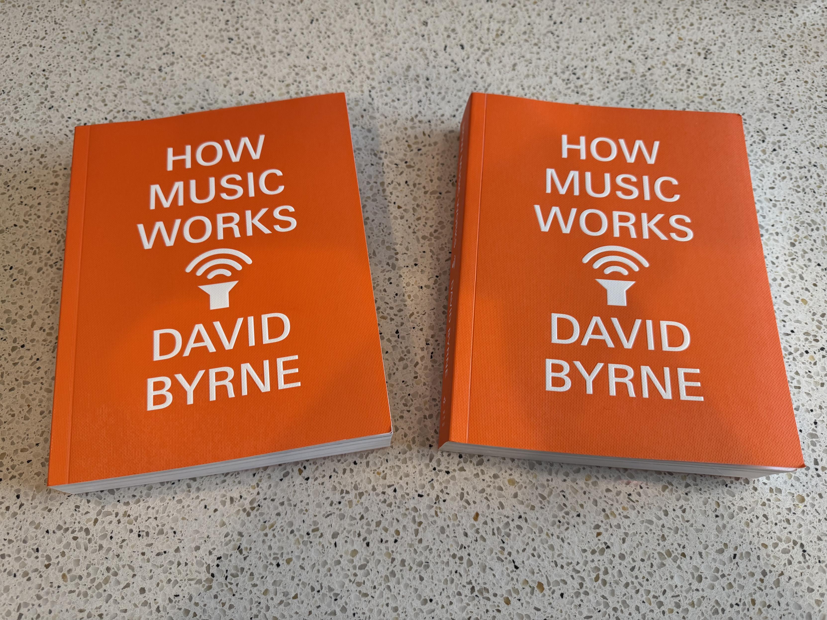 Two identical paperbacks of David Byrne’s “How Music Works”, which is bright orange with all caps grotesque title over author’s name, one word per line, with very simple sign for a speaker radiating music up at the title between the two.the books are both laying face up on my kitchen counter.HOWMUSICWORKSDAVIDBYRNE.