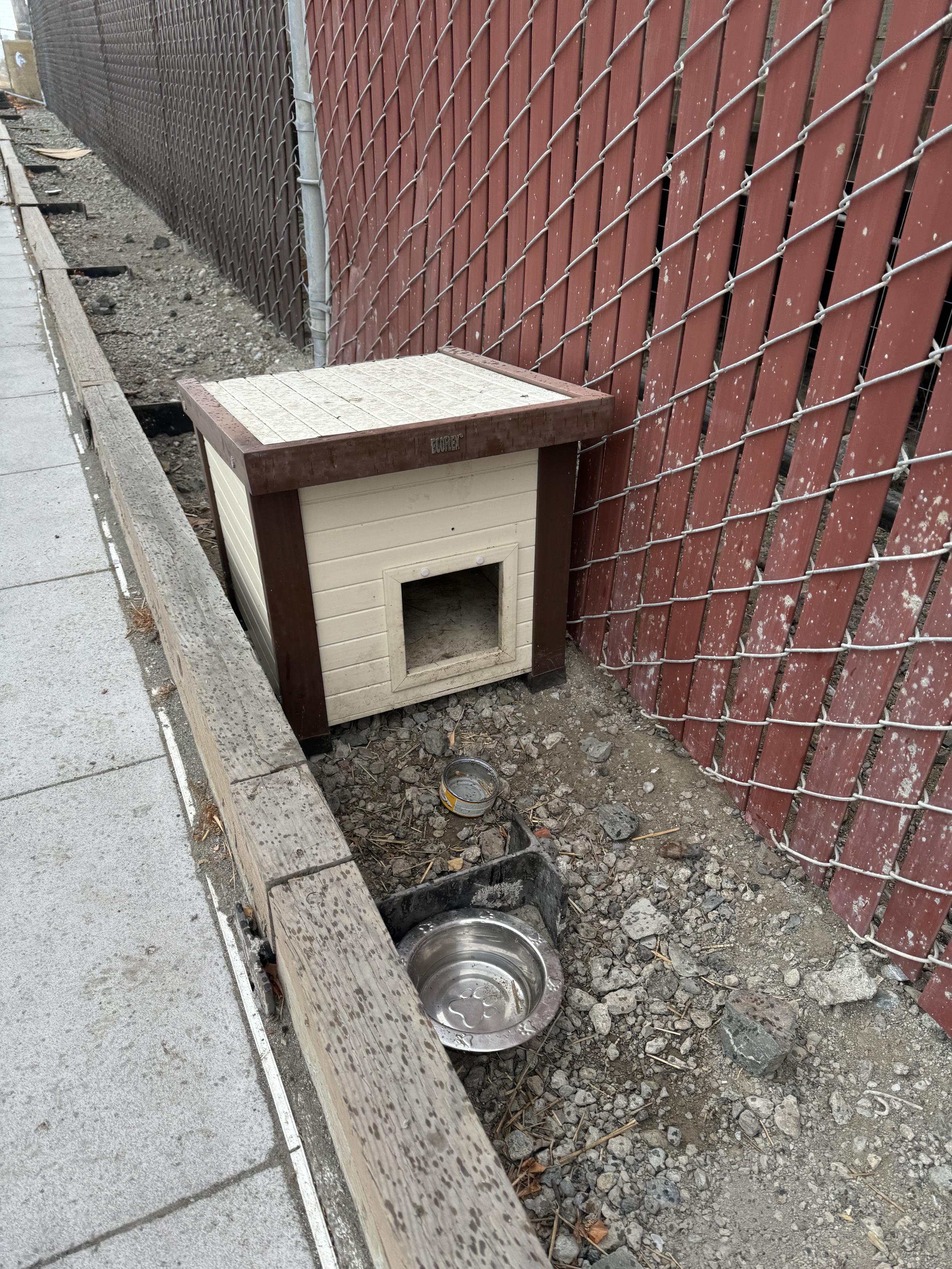 a closer photograph of one of the cat houses situated between a sidewalk curb and a chain link fence with narrow panels slotted through the fencing, confirmed to be a cat house by the open empty can of cat food in front of it next to an empty water bowl