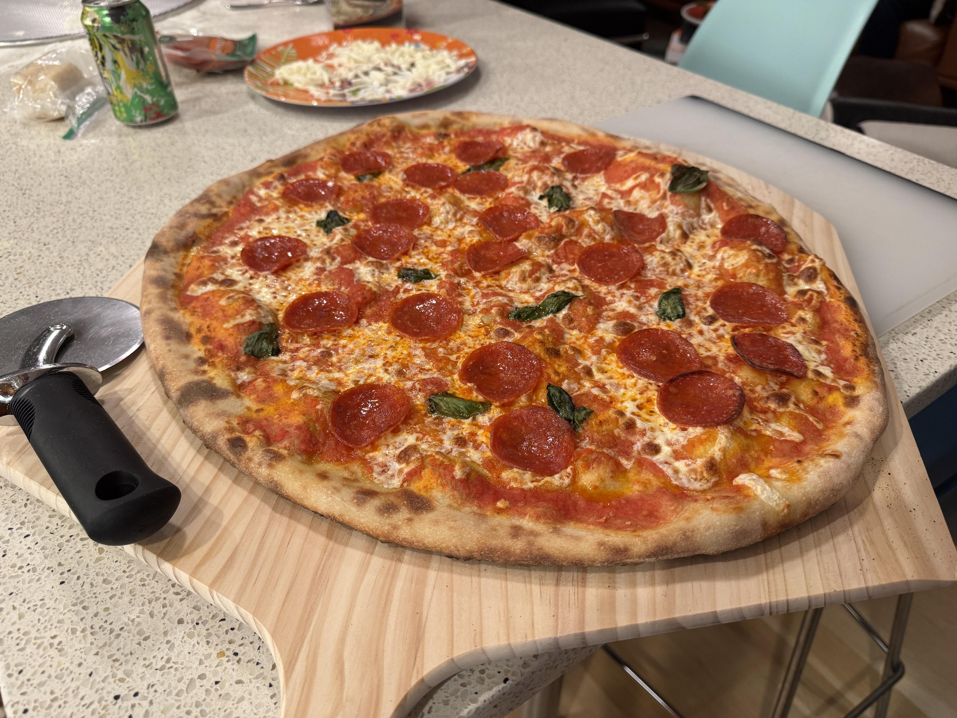 photograph, finished extra large new york style pizza with sauce, cheese, basil, and pepperoni on a wood peel on a counter, a pizza wheel is laying next to the pizza on the left