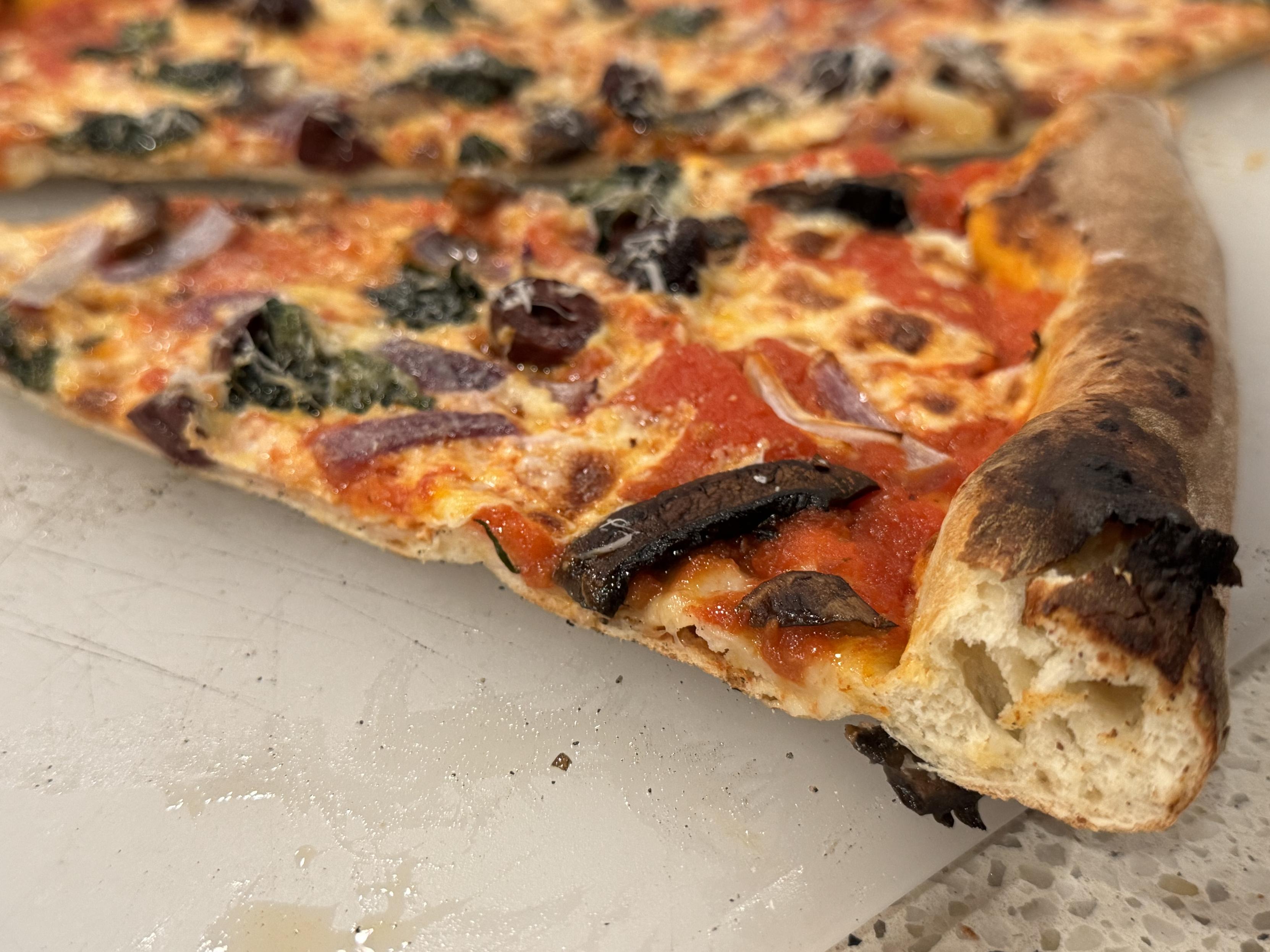 photo, a slice of pizza on a cutting board.the pizza is topped with smoked kalamata olives (holy moly, amazing), red onion, mushroom, basil, pecorino, mozz.the sauce: drain a can of whole peeled san marzanos, dicard the liquid, and zap what’s left in the blender w/ one head of garlic and generous pinches of salt, sugar, and oregano.