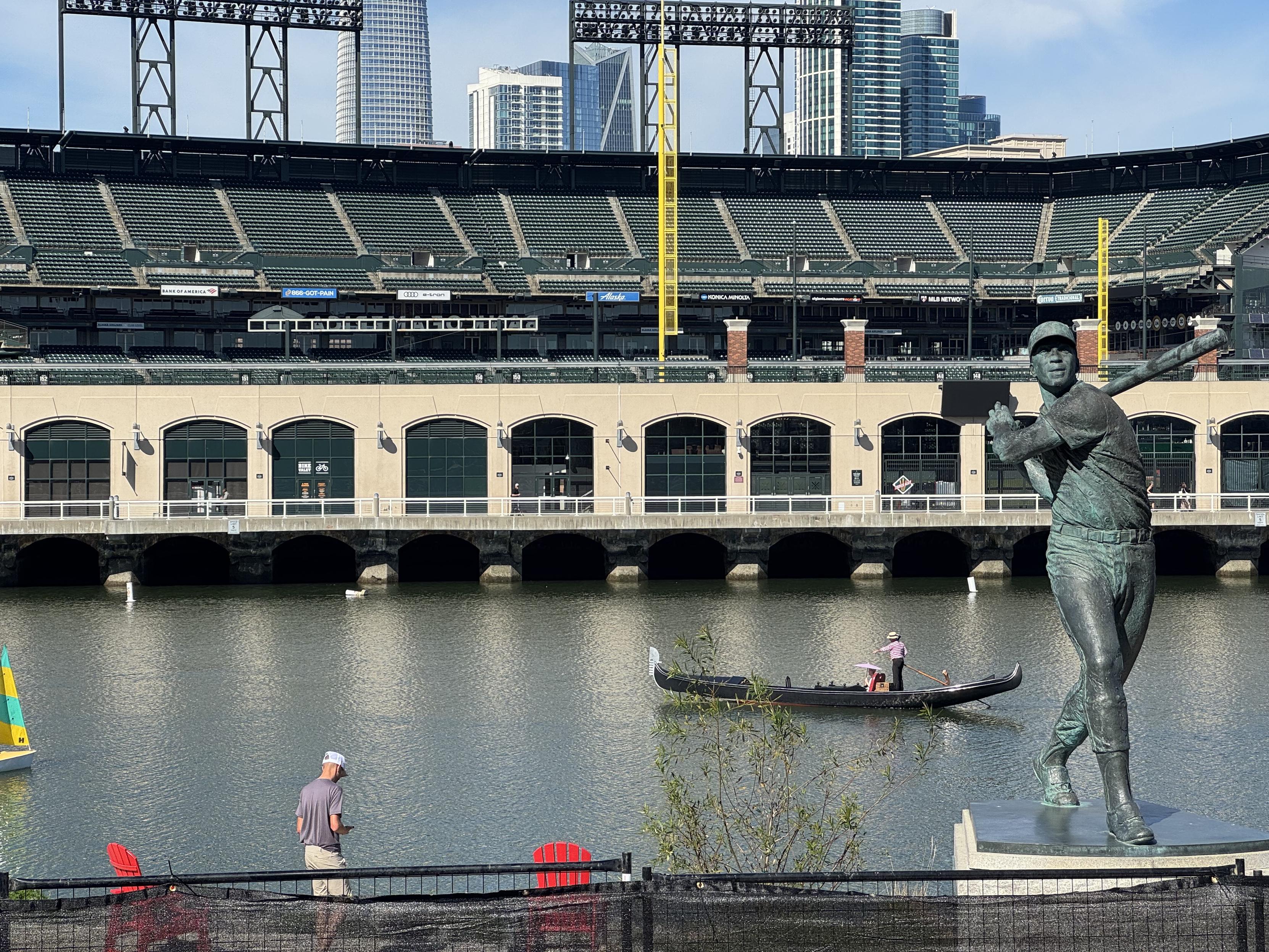 photograph of the san francisco giants stadium from beyond right field and across a body of water. in the foreground, a twenty foot statue of baseball player who just swing their bat is on a small pedestal, but what its eyes catching is that in the water, a gondola is being oared by a gondolier wearing a red and white striped shirt and a flat brimmed straw hat. the sole occupant of the umbrellas is holding a paper straw for the afternoon sun. i've never been to Venice but i assume these people have or wish they had.