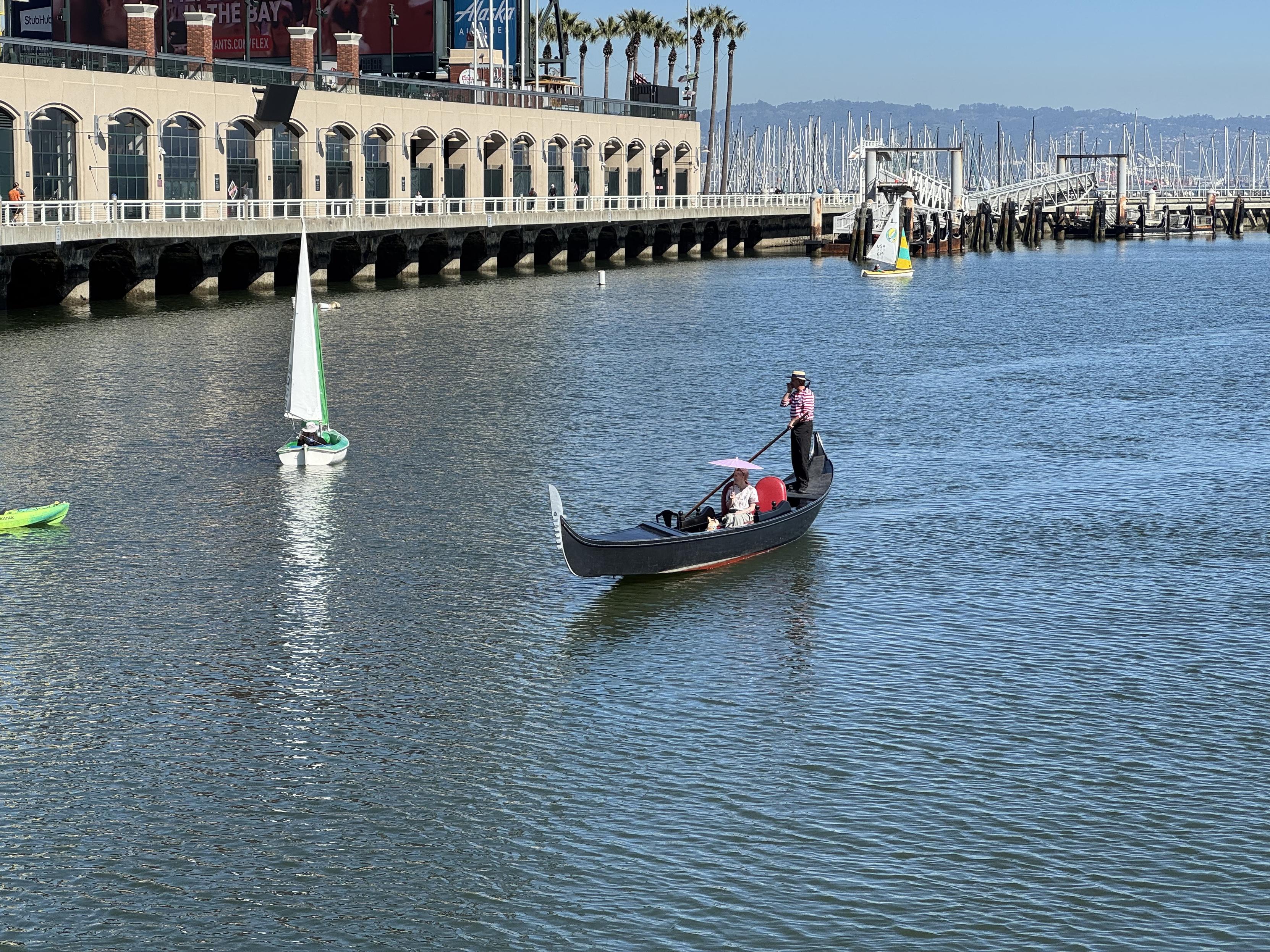 photograph of the san francisco giants stadium the bridge the gondolier is about to go under. a gondola is being oared by a gondolier wearing a red and white striped shirt and a flat brimmed straw hat. the sole occupant of the umbrellas is holding a paper straw for the afternoon sun. i've never been to Venice but i assume these people have or wish they had. there are a few other small sailboats around them.