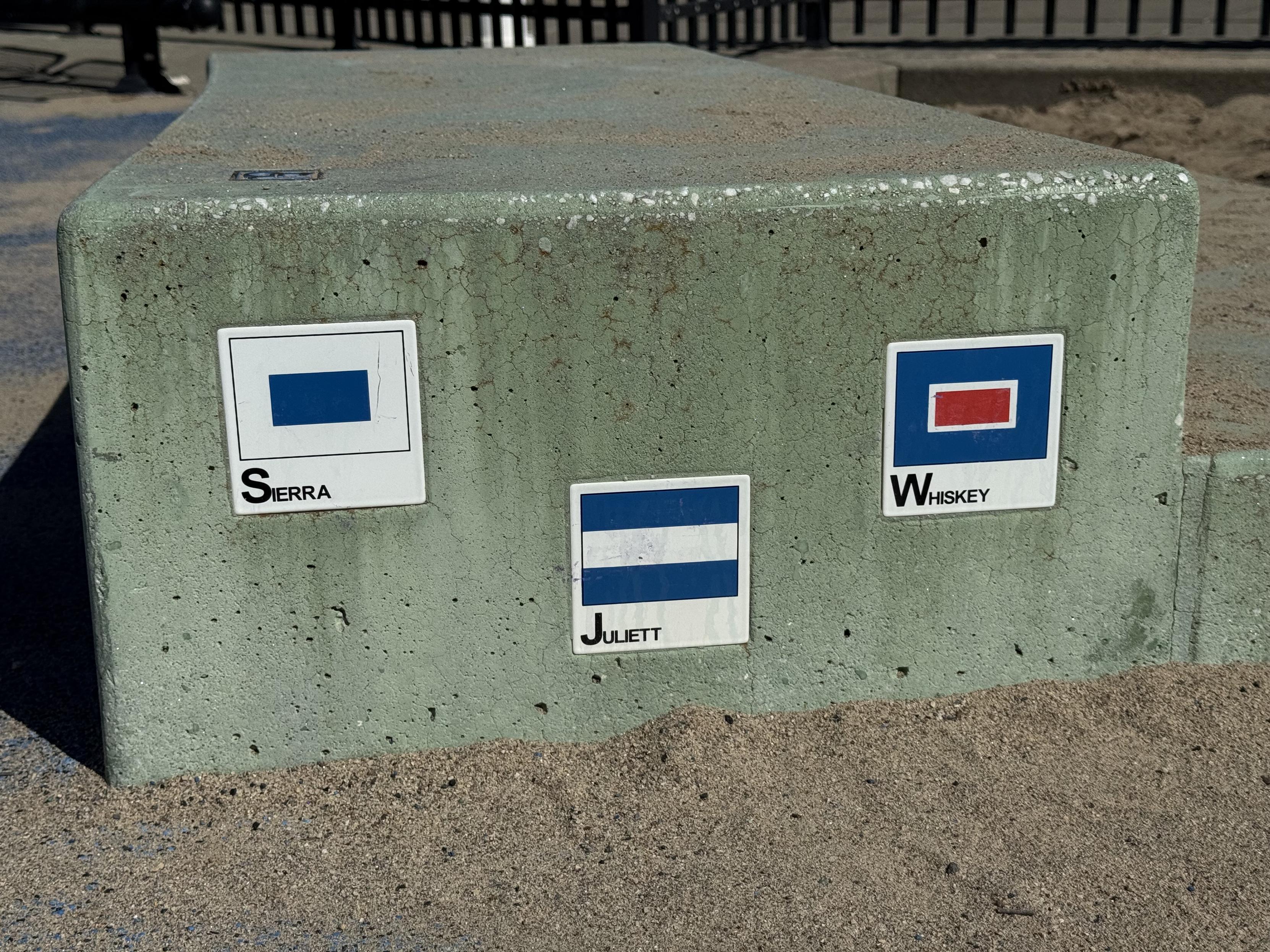 photograph of small nautical flag (i assume?)  tiles embedded in concrete steps at a toddler playground in san francisco with Sierra, Juliett, and Whiskey written under each flag, The initial letters are large while the remainder are small caps.