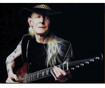 a photo of johnny winter in a black sleeveless t-shirt and a black cowboy hat holding a burst gibson firebird over a black background.