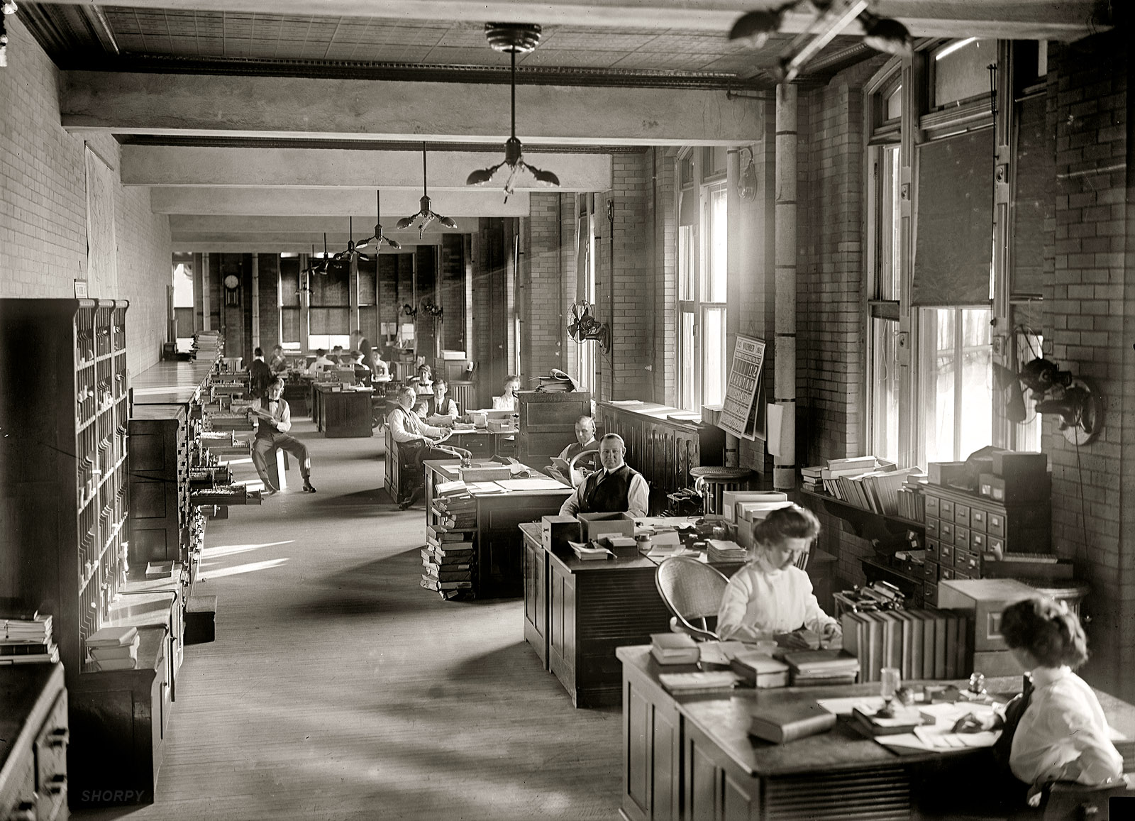 November 1912. "Government Printing Office, Washington." - black and white photograph of a the interior of an early 20th century office setting, with half a dozen desks along brick walls to the right lit by large, tall windows. At each is seated a man or woman in period dress. Along the left the entire wall is cabinets, most of which have a drawer open.